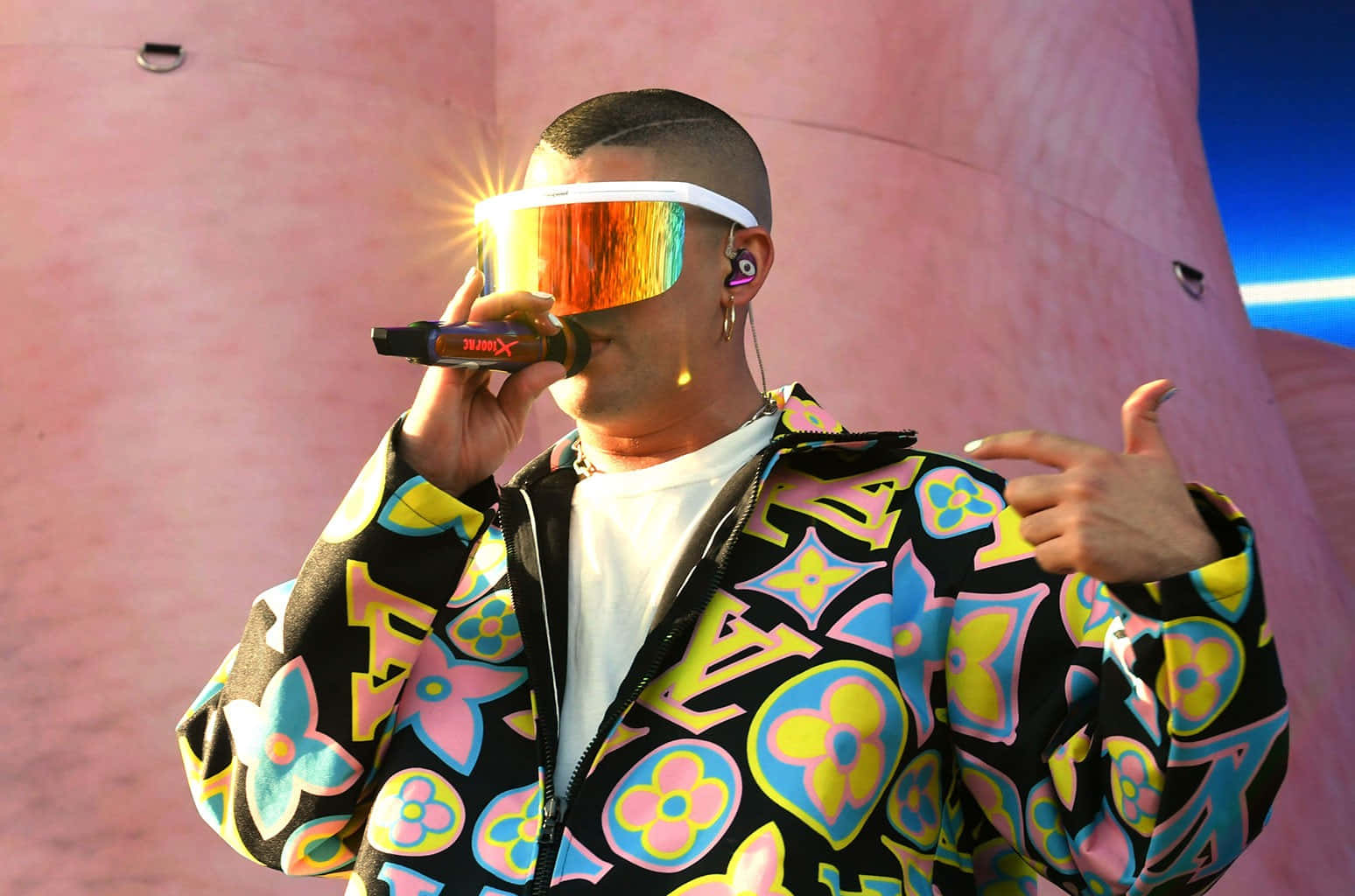Bad Bunny 桌布 壁紙 — 一位身穿色彩繽紛花卉外套和反光護目鏡的表演者，在粉紅色弧形牆前對著麥克風唱歌。