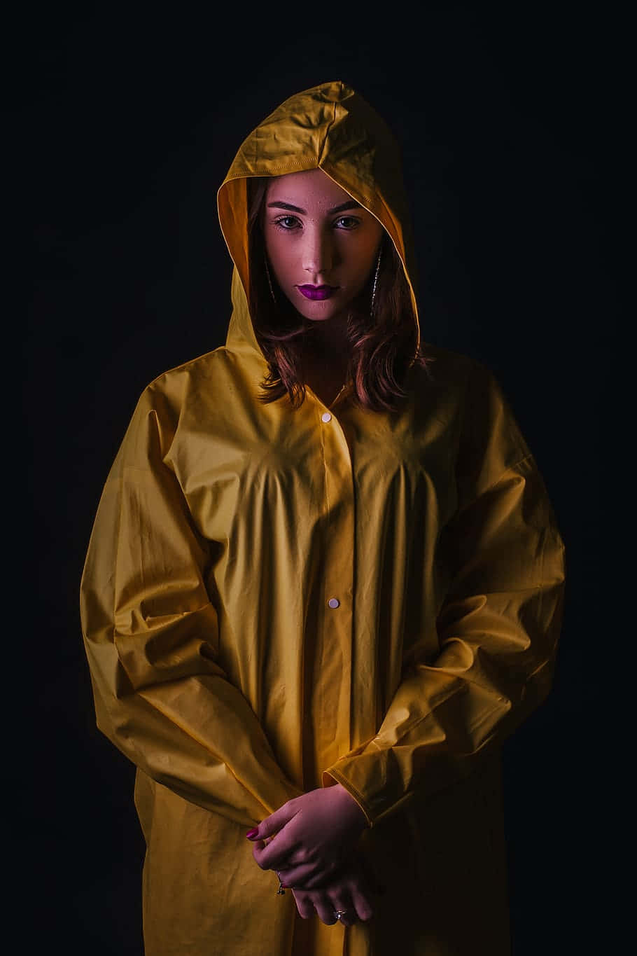 Yellow Raincoat 桌布 壁紙 — 一位身穿芥末黃色雨衣的女性，在黑色背景前，帶著強烈的眼神和紫色唇膏站立。