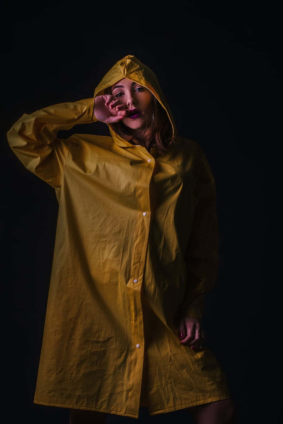 Yellow Raincoat 桌布 壁紙 — 一位身穿芥末黃雨衣的女性站在黑色背景前，手舉到臉部附近，擺出戲劇性的姿勢。