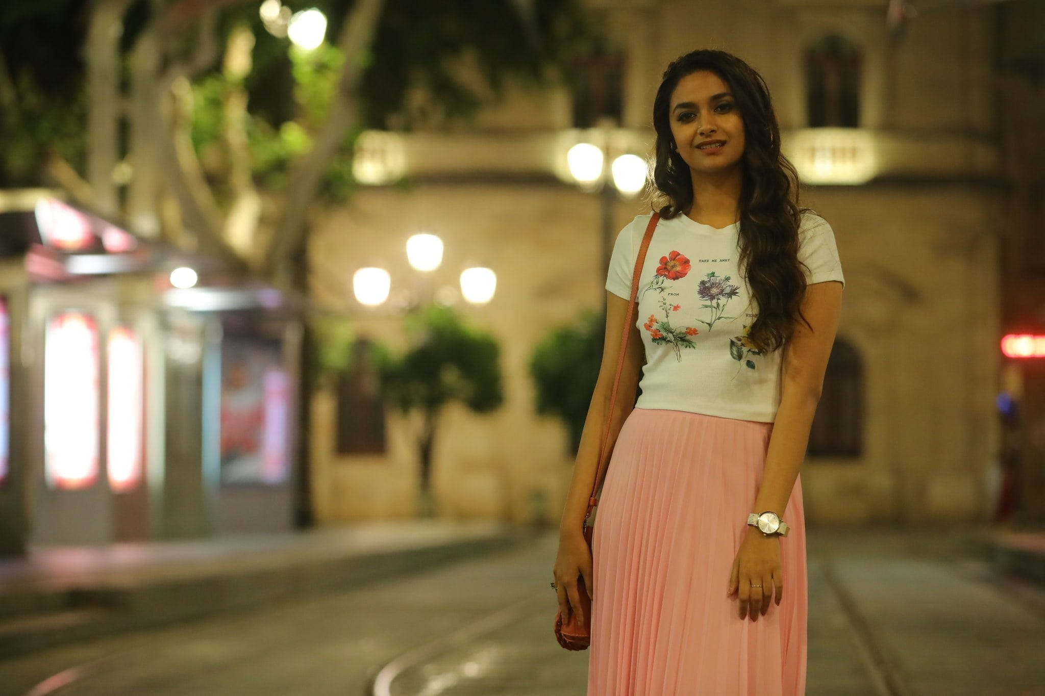 Tamil Actors HD 桌布 壁紙 — 一位身穿花卉T恤和粉紅色百褶裙的年輕女性，在夜晚溫暖燈光的城市街道上輕柔微笑。