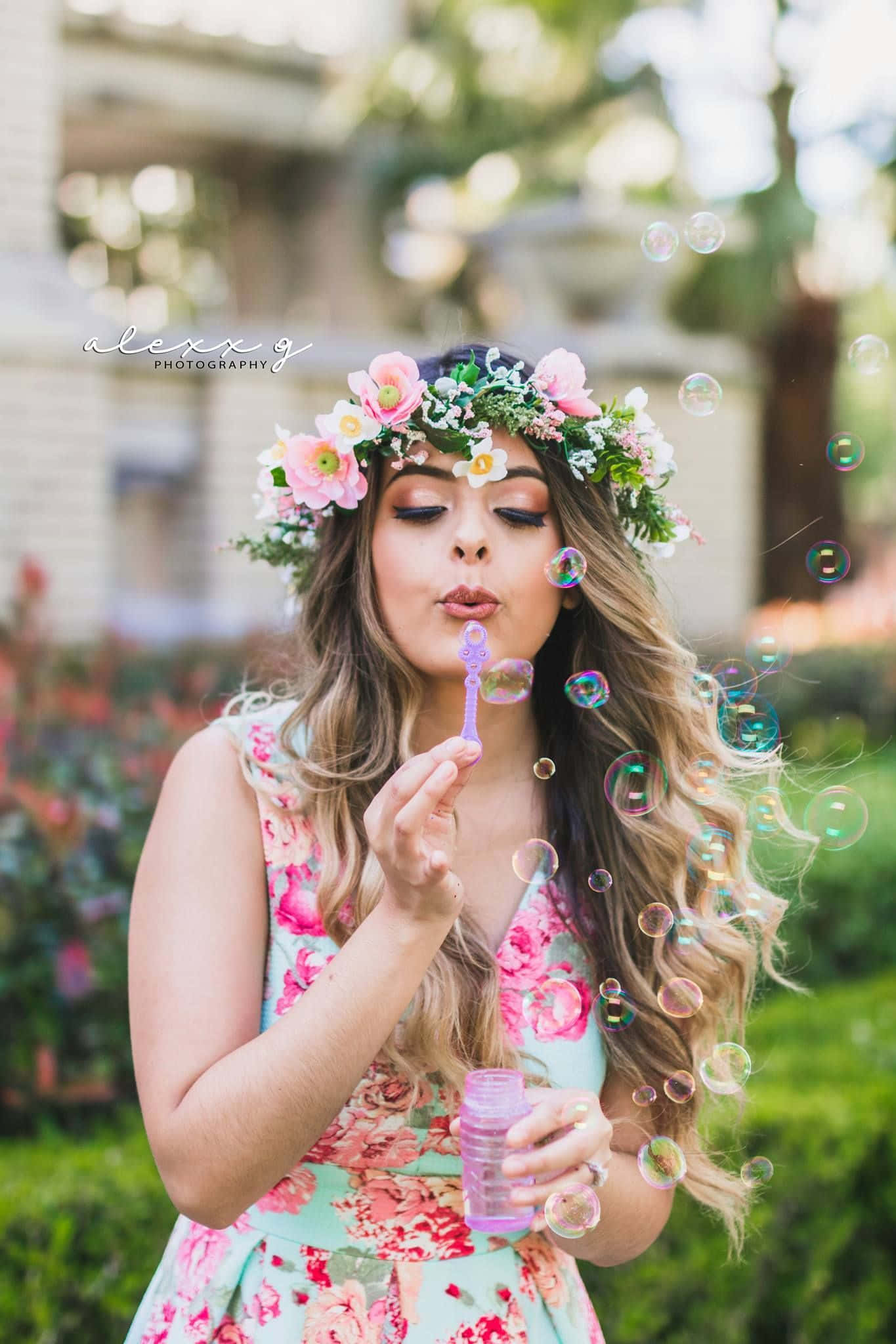 Floral Crown 桌布 壁紙 — 一位身穿花卉連衣裙、頭戴皇冠的年輕女子，在陽光普照的花園中吹出色彩繽紛的泡泡，背景帶有柔和散景效果。