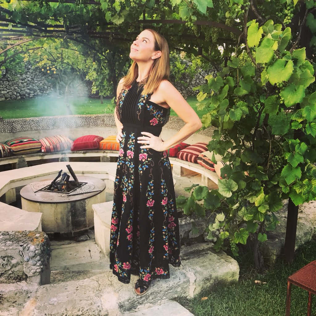 Kate Ritchie 桌布 壁紙 — 一位身穿花卉連衣裙的女性站在黃昏時分茂密花園中一個被藤蔓覆蓋的石頭火坑旁。