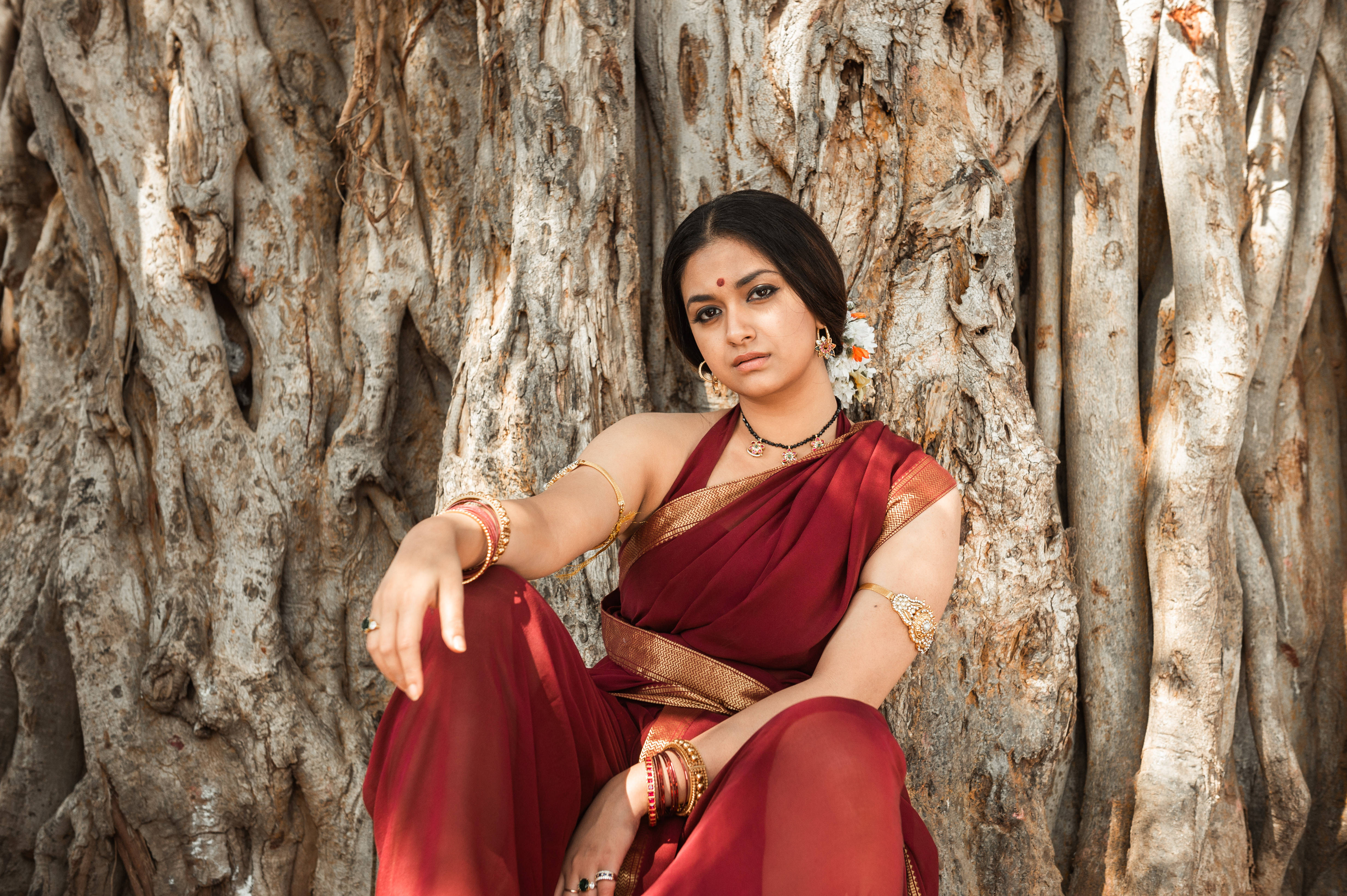 Indian 桌布 壁紙 — 一位身穿華麗紅色紗麗、佩戴金飾的女子，優雅地靠在一棵巨大榕樹的紋理樹皮上。