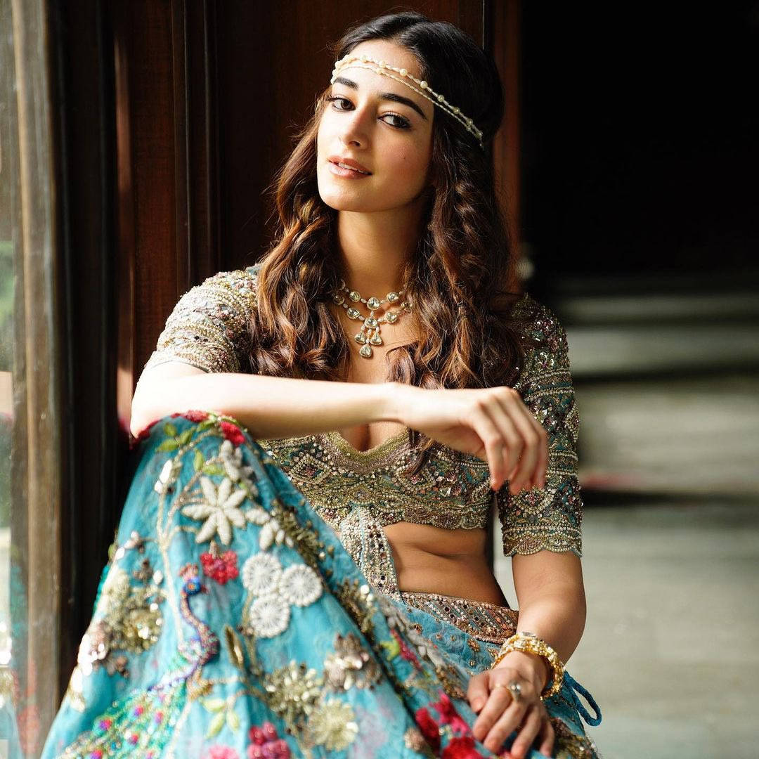 Ananya Pandey 桌布 壁紙 — 一位身穿華麗藍金刺繡長裙（Lehenga）的女性，在柔和自然光沐浴的木門旁優雅地擺姿勢。