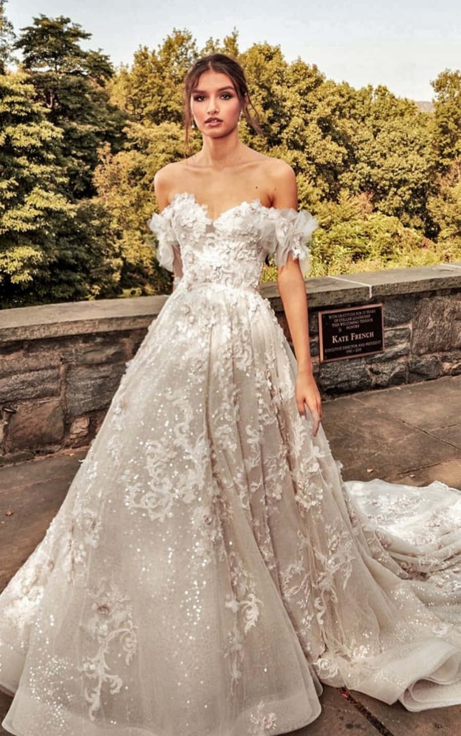 Wedding Dress 桌布 壁紙 — 一位身穿華麗露肩象牙白婚紗的女性，優雅地站在被茂密綠樹環繞的石砌陽台上。