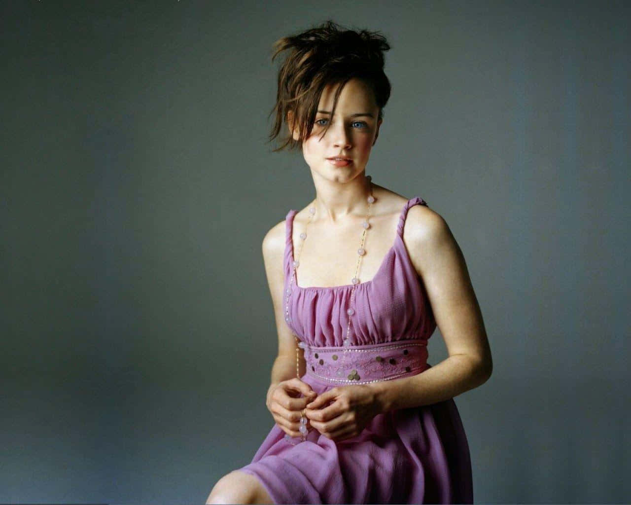 Alexis Bledel 桌布 壁紙 — 一位身穿薰衣草色連衣裙的女性，坐在柔和的灰色背景前，頭髮蓬鬆，目光直視。