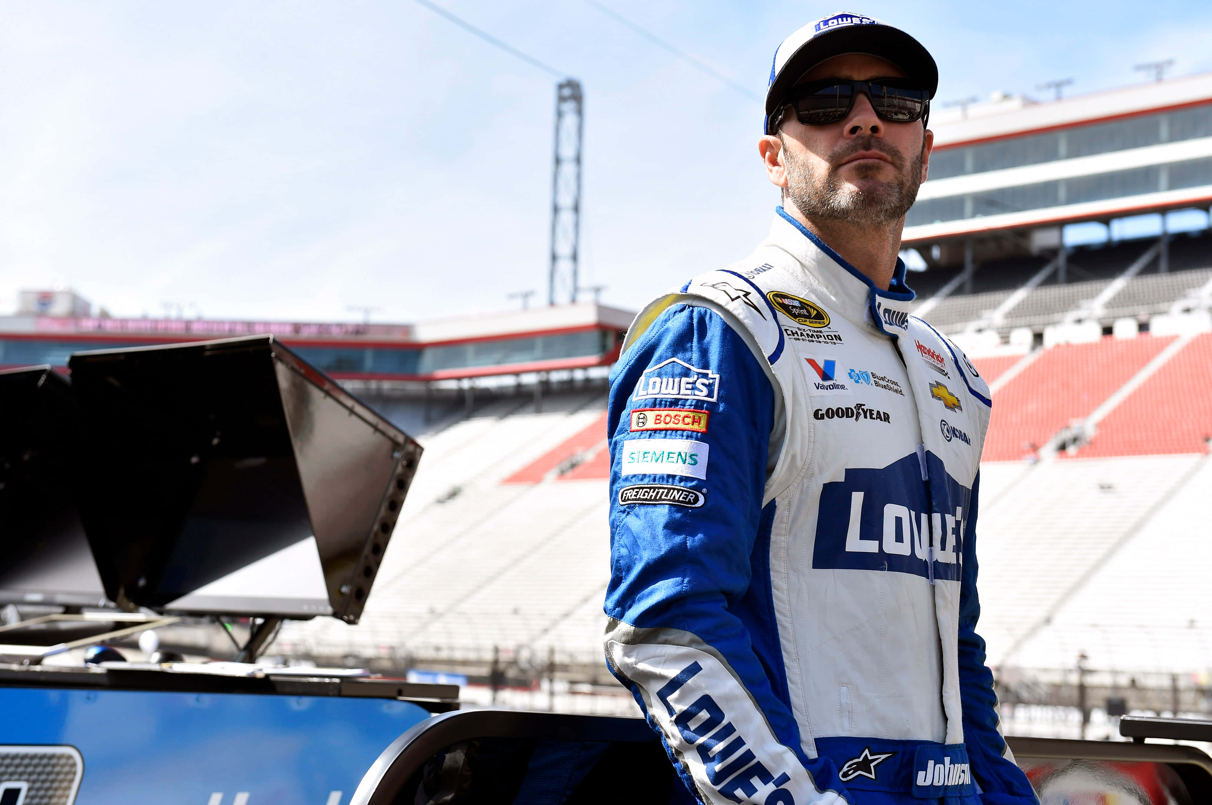 Jimmie Johnson 桌布 壁紙 — 一位身穿藍白Lowe's賽車服的NASCAR車手自信地站在陽光普照的體育場內他的賽車卡車旁。