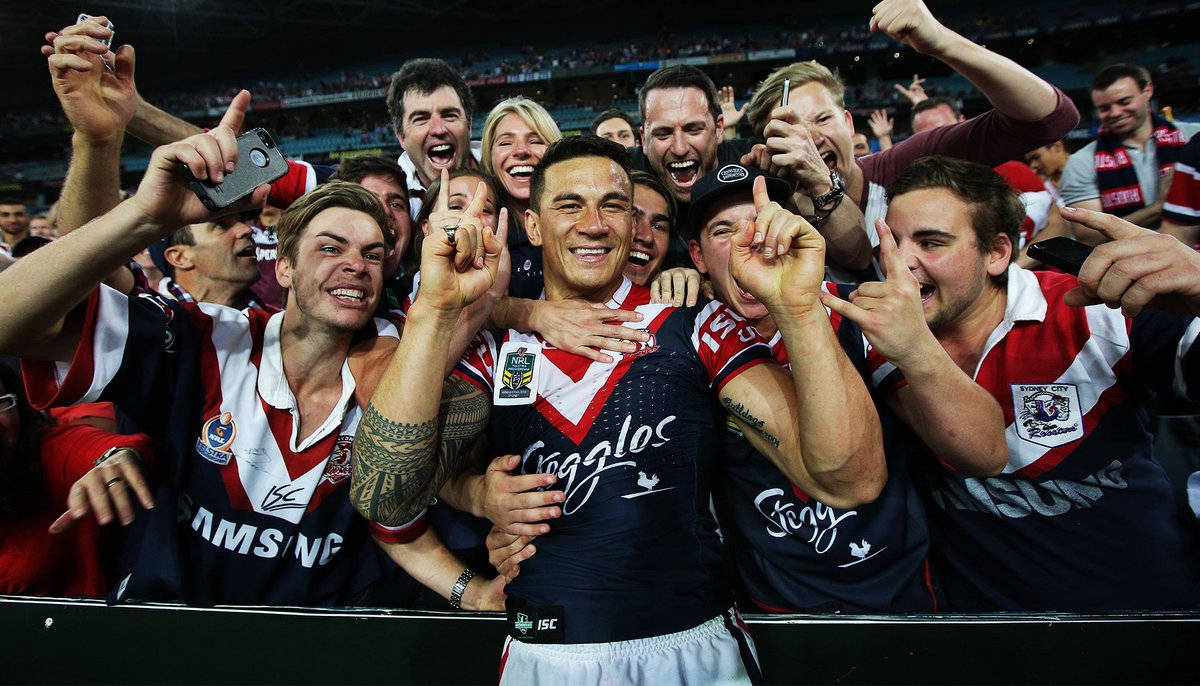 Sonny Williams NRL 壁紙 — 一位身穿藍紅球衣的歡欣欖球聯賽球員在體育場內被歡呼的隊友和球迷擁抱。