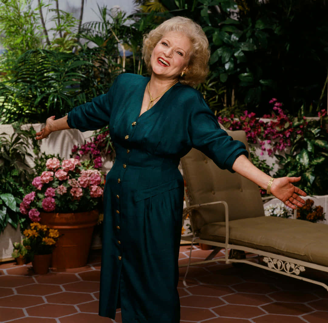 Betty White 桌布 壁紙 — 一位身穿藍綠色連衣裙的快樂女性在開滿粉紅色和黃色花朵的茂盛花園中張開雙臂大笑。