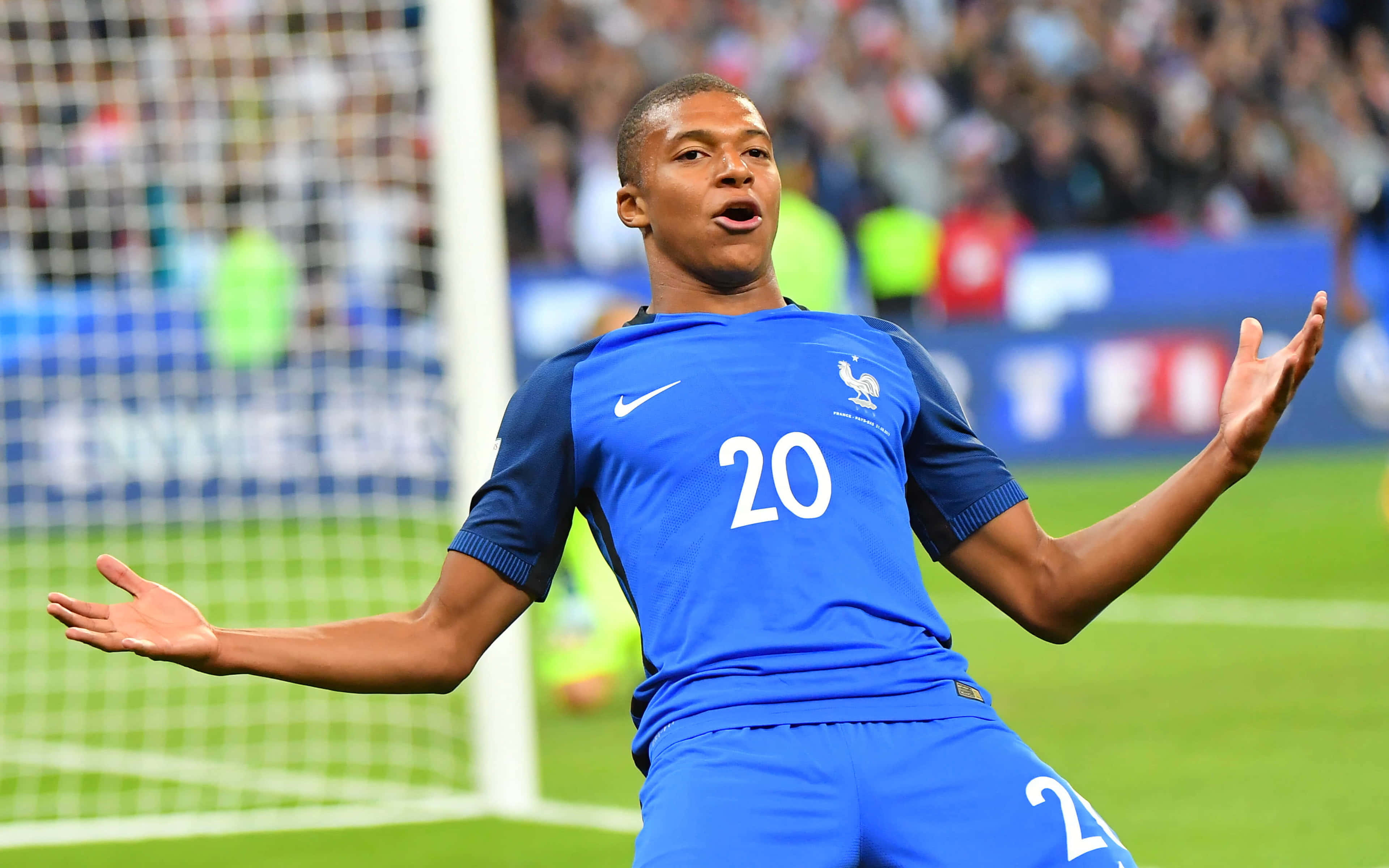 Mbappe 桌布 壁紙 — 一位身穿藍色20號球衣的法國足球員在充滿活力的綠色球場上張開雙臂慶祝。