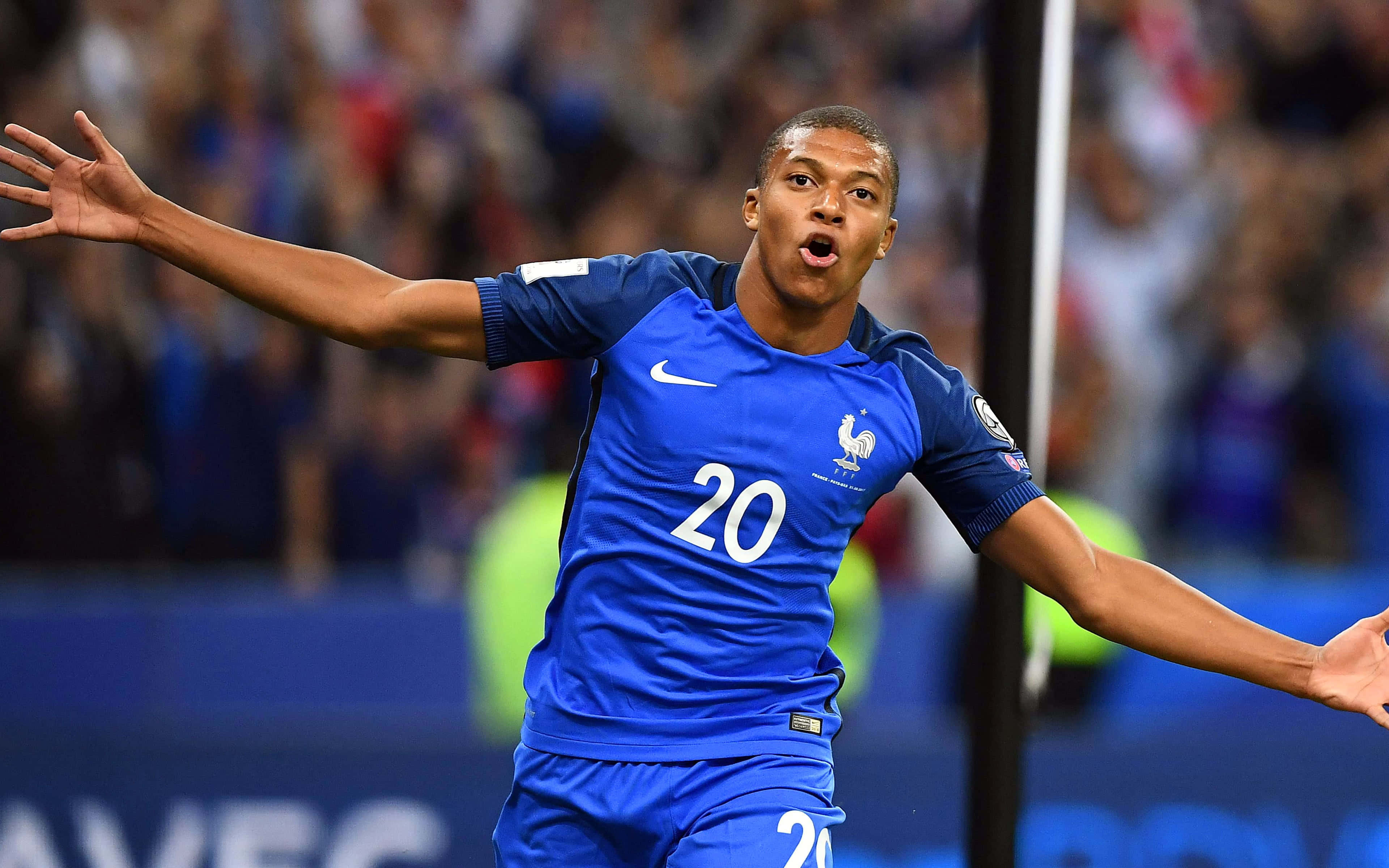Mbappe 桌布 壁紙 — 一位身穿藍色20號球衣的法國足球員在球場上張開雙臂慶祝。