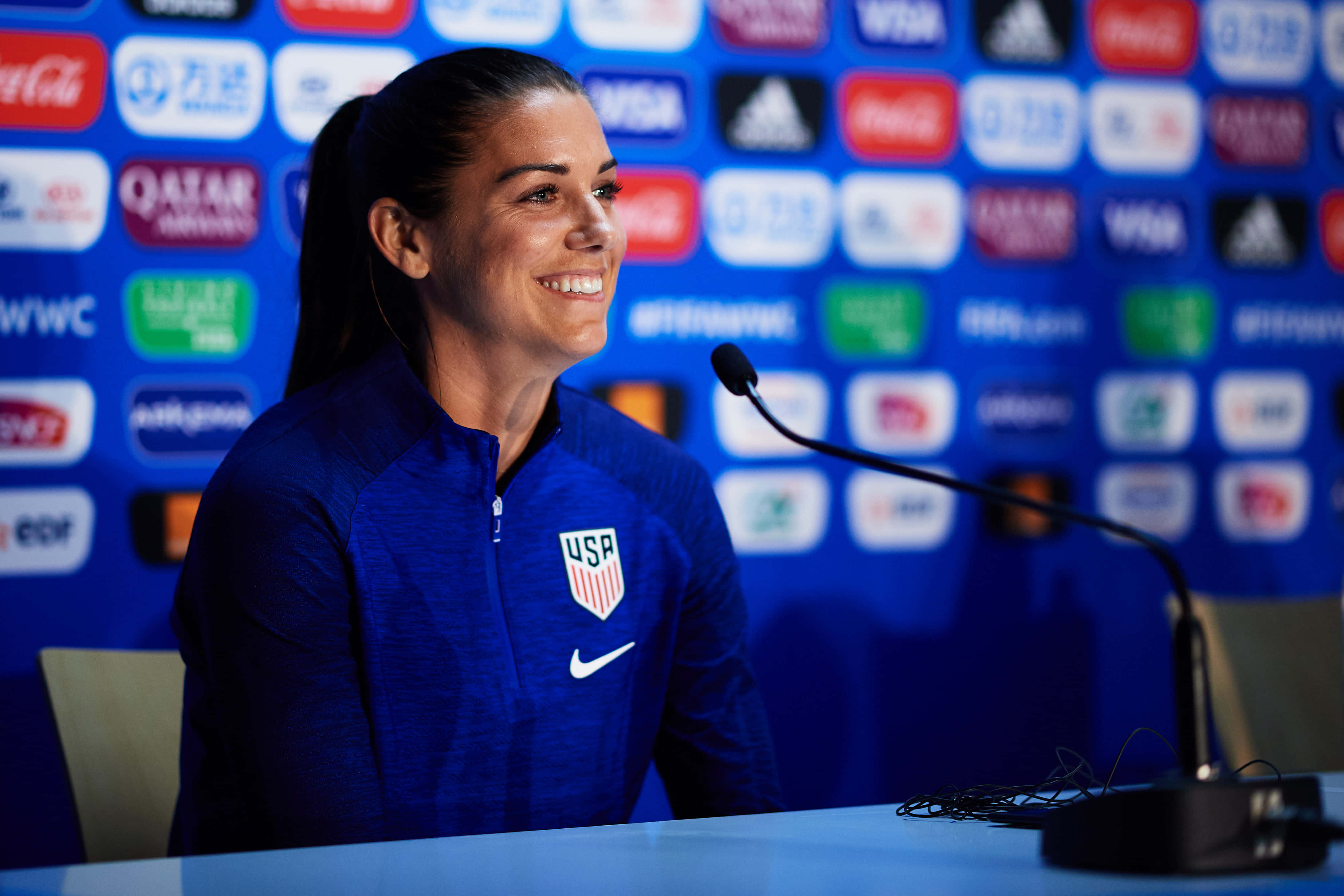 Alex Morgan 桌布 壁紙 — 一位身穿藍色USP球衣、面帶微笑的女足球員坐在記者會的桌子前，身後有咪高峰和贊助商標誌。
