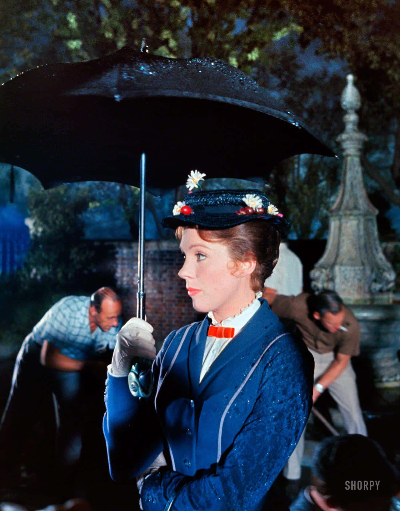 mary poppins 桌布 壁紙 — 一位身穿藍色外套、戴著花卉帽子的女士在雨中撐著黑色雨傘，背景有工人。