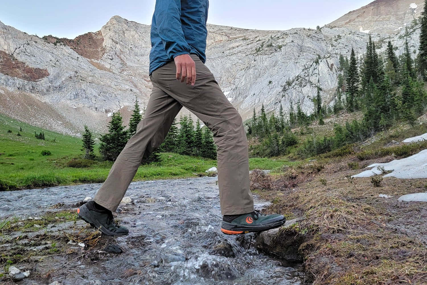 Hiking 桌布 壁紙 — 一位身穿藍色外套和棕色褲子的遠足者，踏過一條岩石山間溪流，背景有松樹和雪斑。