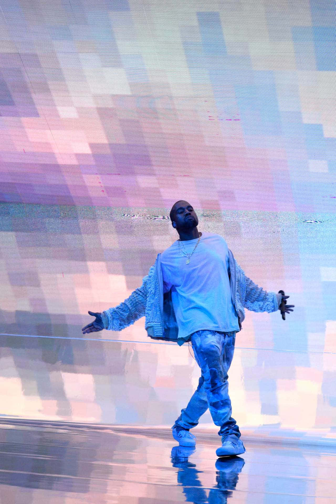 Kanye 桌布 壁紙 — 一位身穿藍色服裝的表演者站在反射舞台上，雙臂伸展，背景是充滿活力的像素化背景。