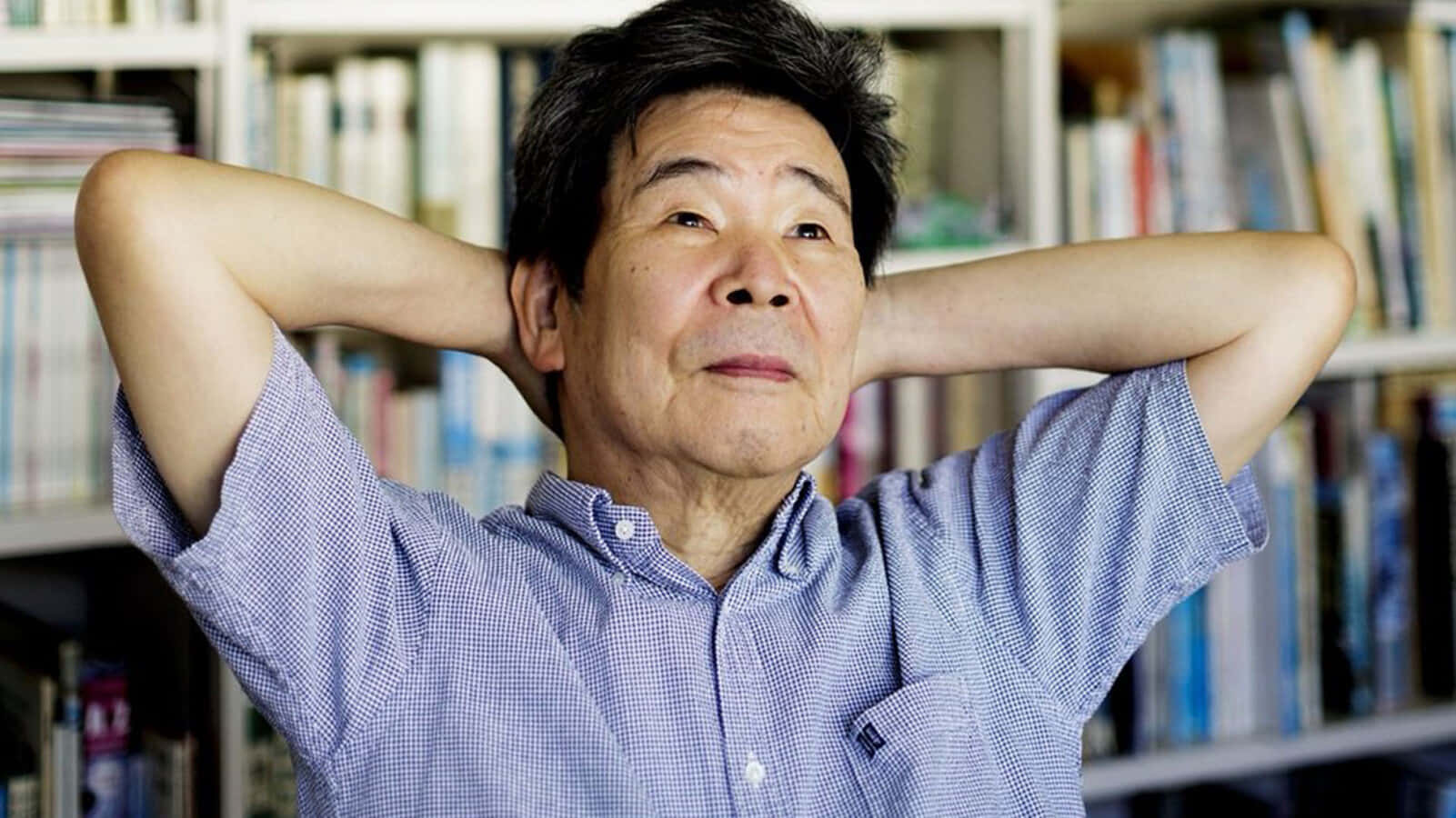 Isao Takahata 桌布 壁紙 — 一位身穿藍色格仔恤衫的長者，在放滿書架的房間裡將雙臂枕在腦後休息。