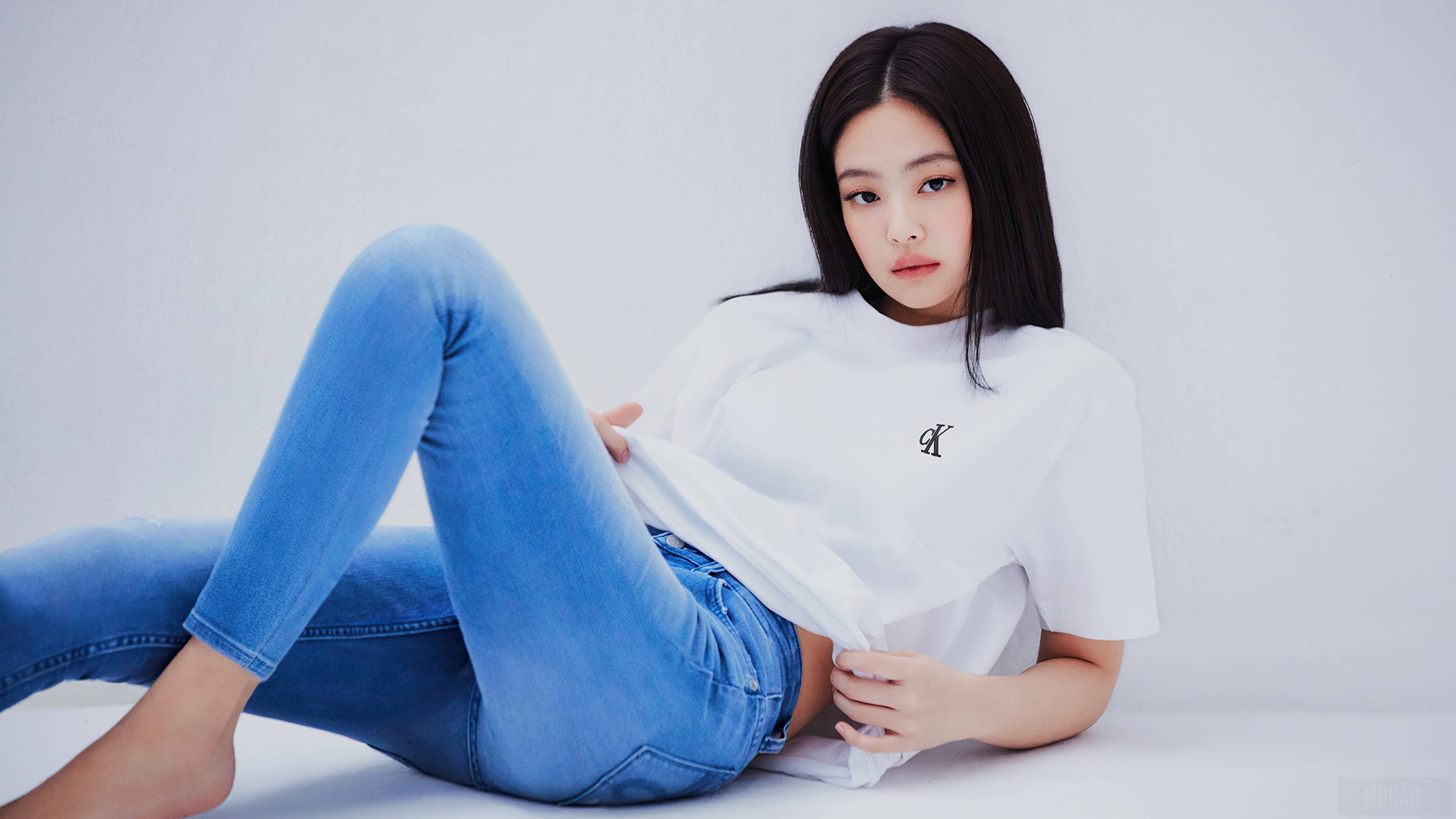 Jennie Kim Calvin Klein 壁紙 — 一位身穿藍色牛仔褲和白色T恤的年輕女士在白色地板上躺著，背景是無縫的白色牆壁。