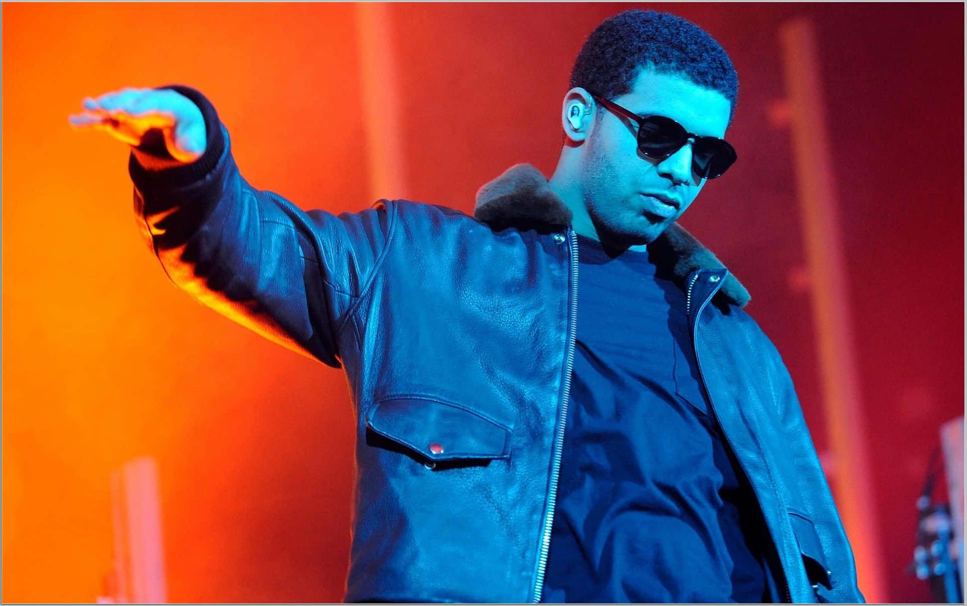 Drake 桌布 壁紙 — 一位身穿藍色皮褸和太陽眼鏡的表演者在充滿活力的橙色和藍色舞台燈光下自信地做手勢。