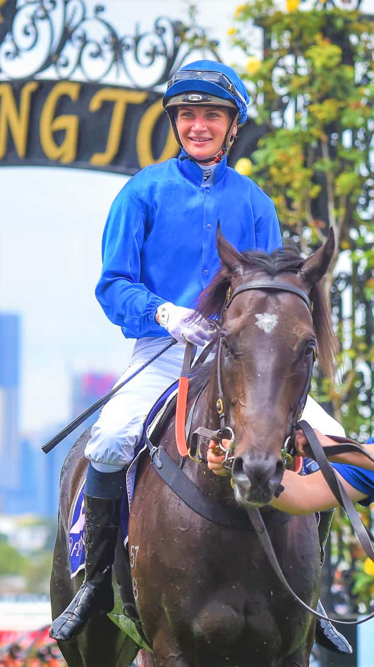Jamie Kah 桌布 壁紙 — 一位身穿藍色絲綢、面帶微笑的騎師，騎著一匹深棕色賽馬，在一個裝飾著綠色樹葉的拱門下。