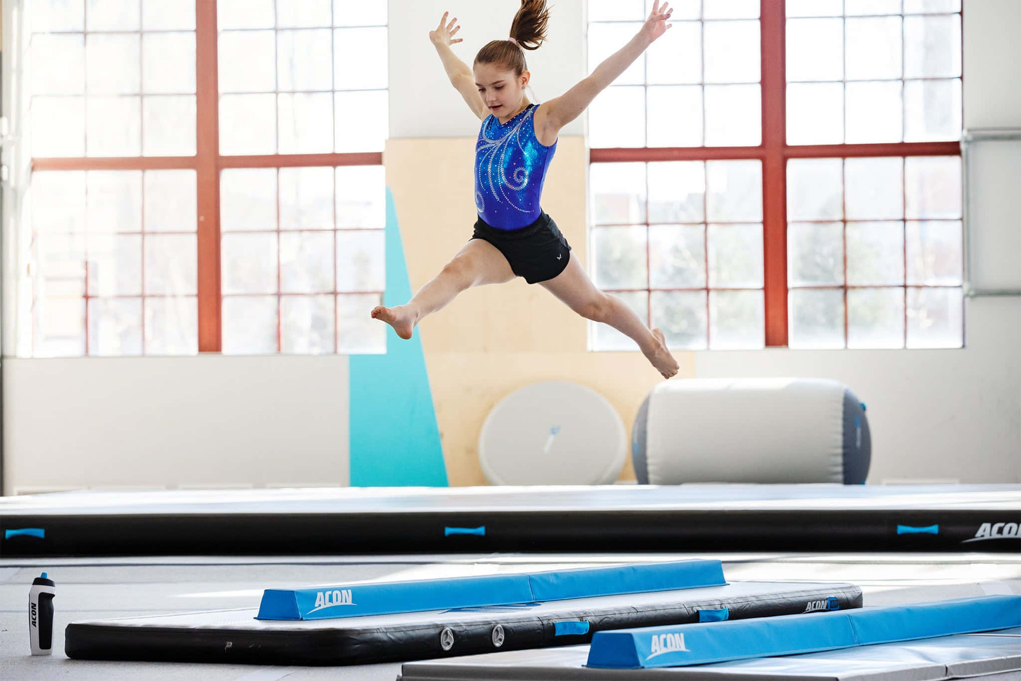 Gymnastics Mat 桌布 壁紙 — 一位身穿藍色緊身衣的年輕體操運動員，雙臂伸展，表情專注，優雅地跳過體操墊。