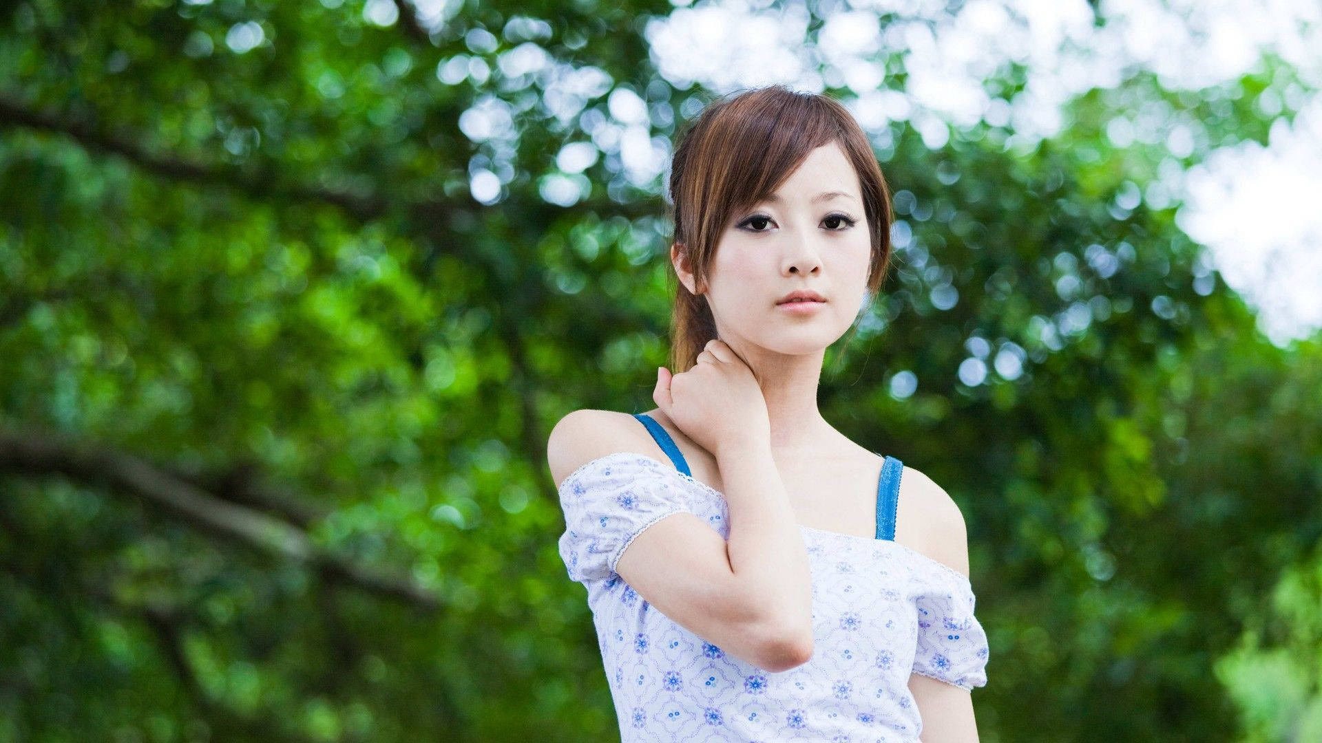 女孩 桌布 壁紙 — 一位身穿藍色花卉上衣的年輕女子優雅地站在鬱鬱蔥蔥的綠樹之間，柔和陽光穿過樹葉灑落。