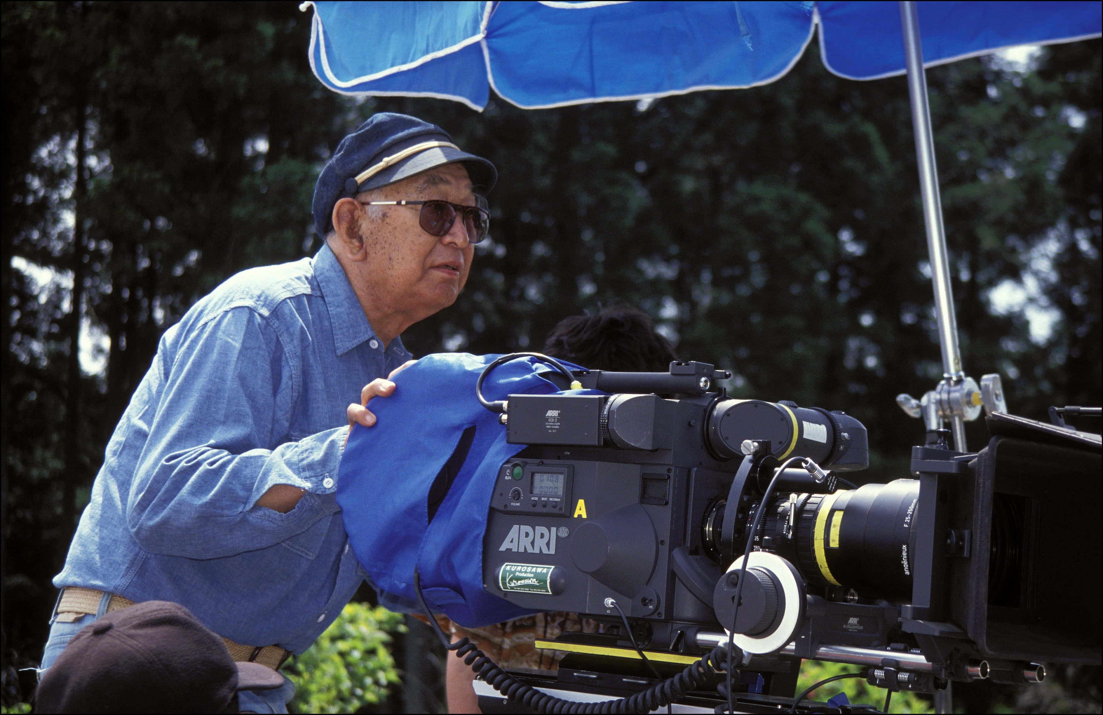 Akira Kurosawa 桌布 壁紙 — 一位身穿藍色襯衫和帽子的年長男性，在森林戶外環境中，於藍色雨傘下操作大型專業電影攝影機。