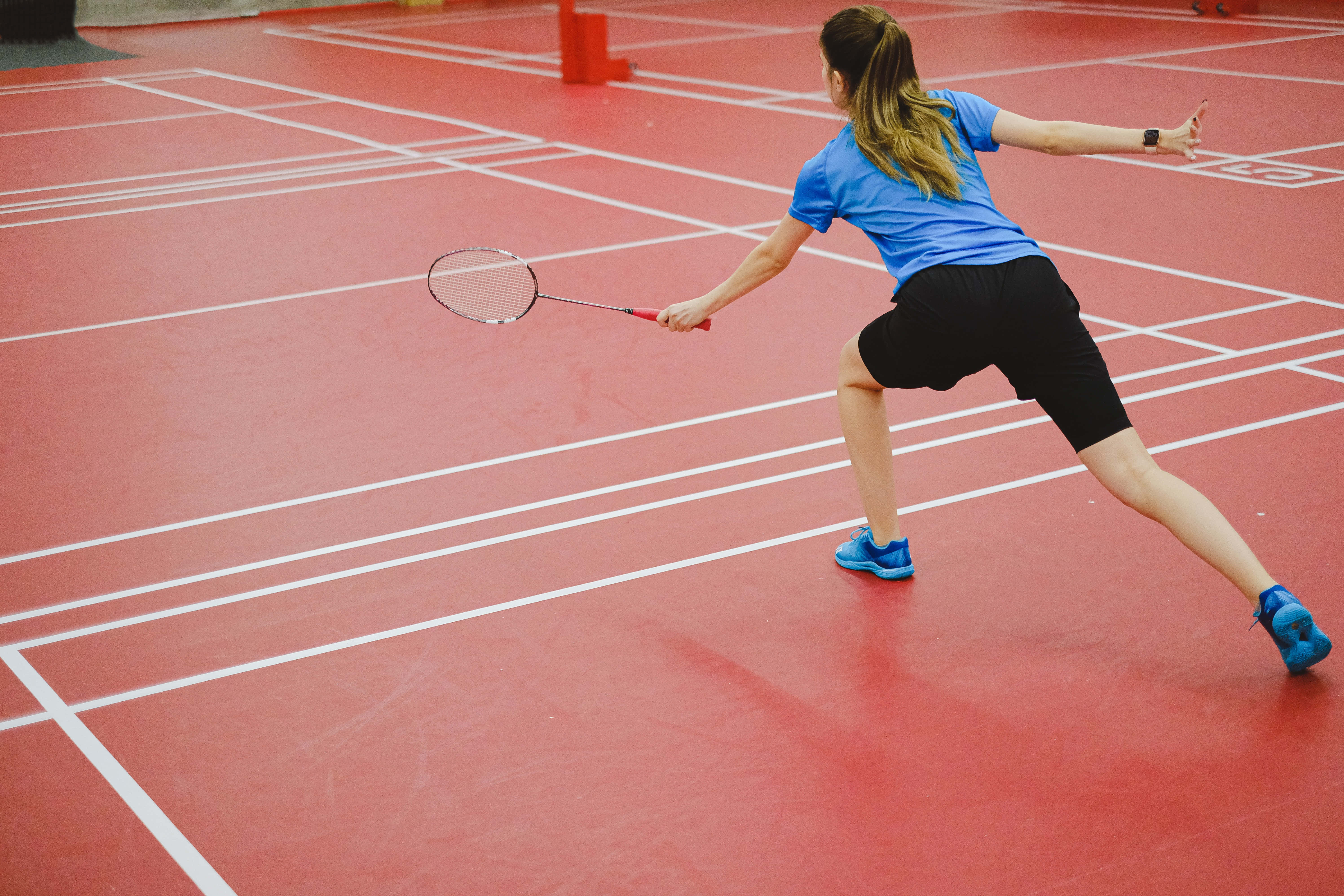 Badminton 桌布 壁紙 — 一位身穿藍色襯衫和黑色短褲的女羽毛球運動員在帶有白色線條的紅色場地上向前衝刺，揮動她的球拍。