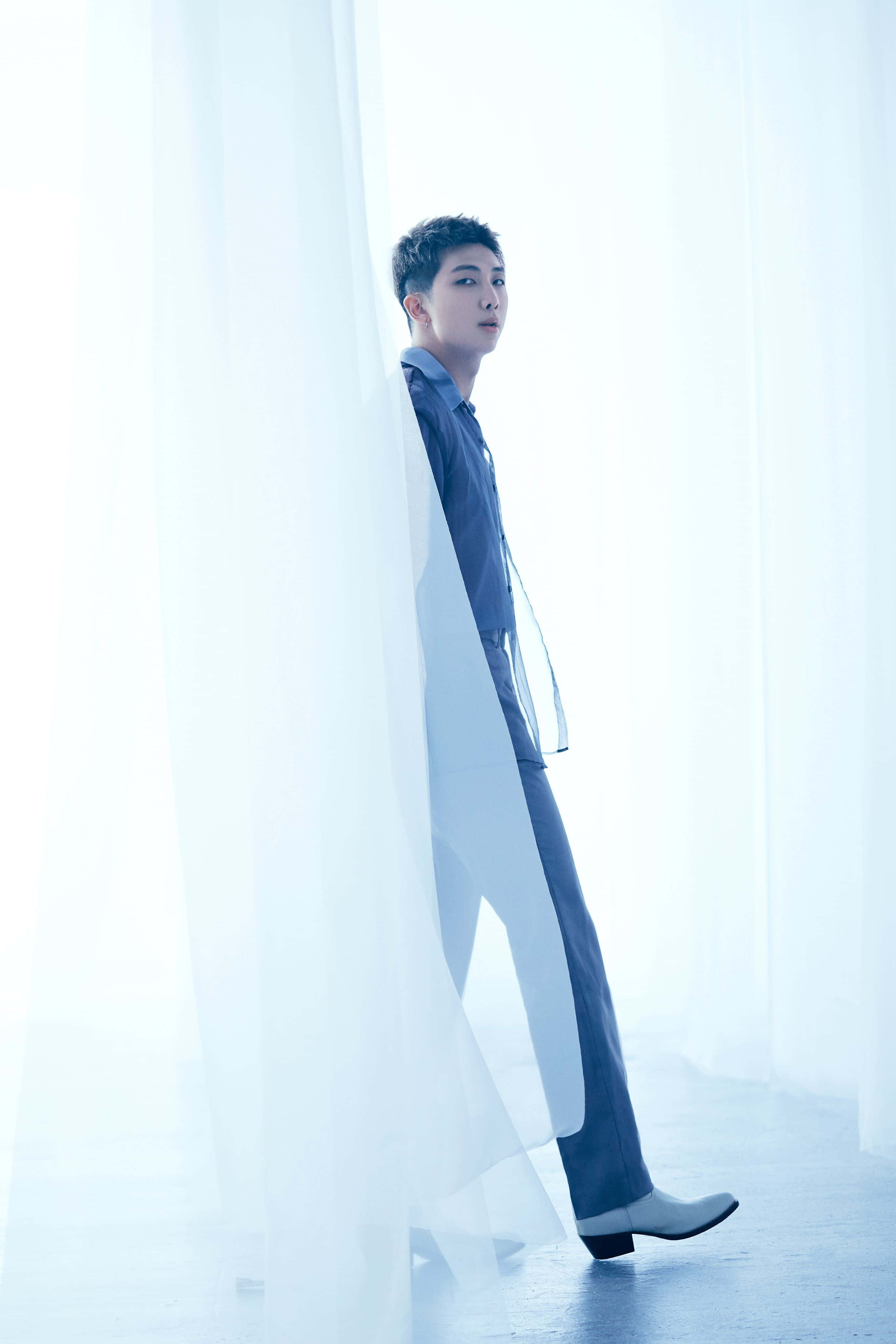 Big Hit Entertainment 桌布 壁紙 — 一位身穿藍色西裝的年輕男子側身站在透明白色窗簾旁，窗簾沐浴在柔和散射的光線中。