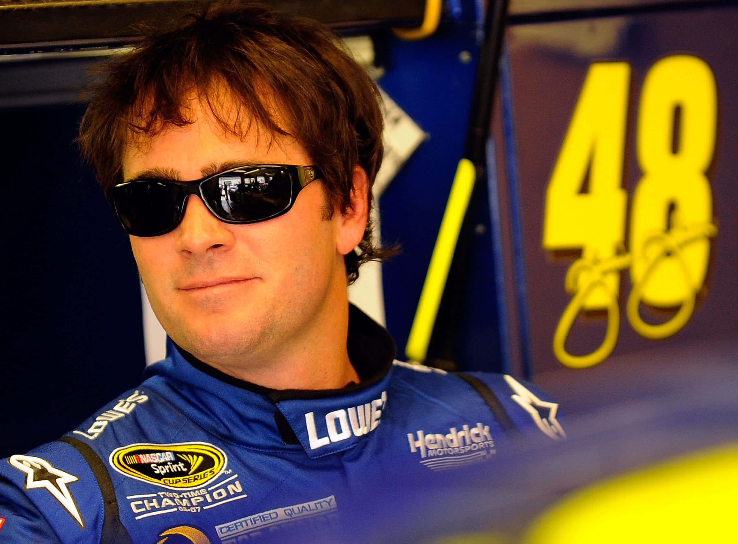Jimmie Johnson 桌布 壁紙 — 一位身穿藍色賽車服和太陽眼鏡的NASCAR車手自信地在他的48號賽車旁擺姿勢。