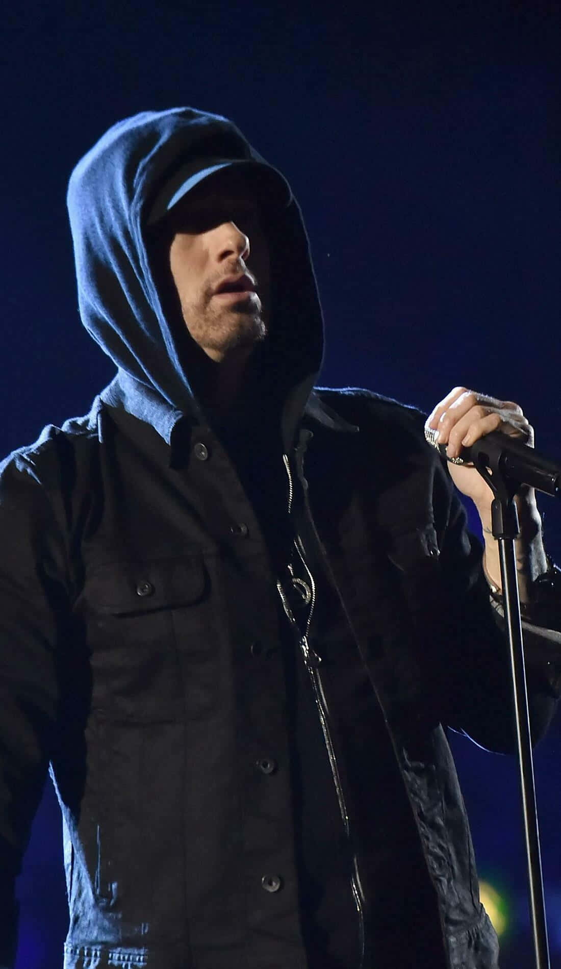Eminem 桌布 壁紙 — 一位身穿藍色連帽衫和黑色外套的男說唱歌手在戲劇性的藍色燈光下握著麥克風在舞台上表演。