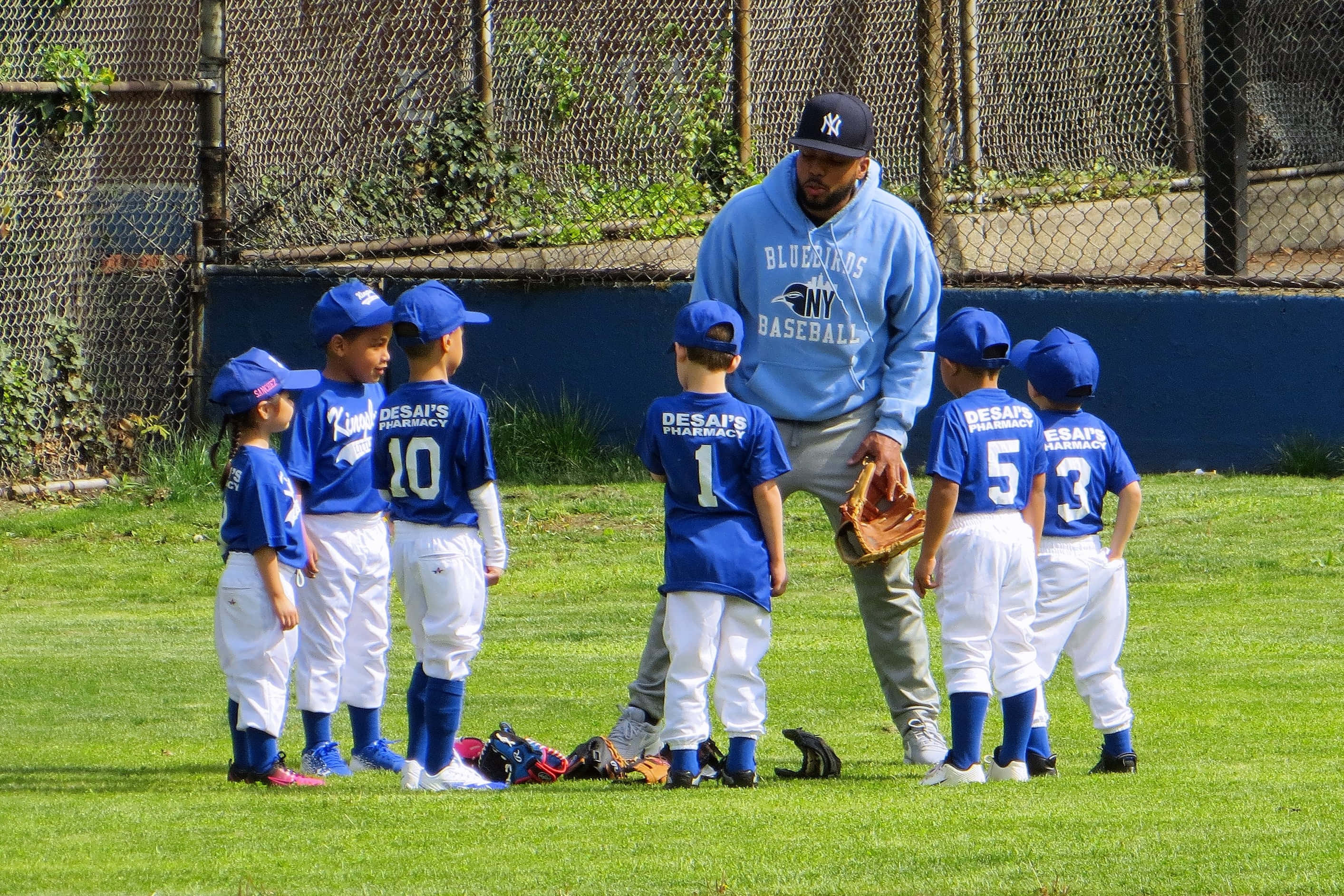 Little League Baseball 桌布 壁紙 — 一位身穿藍色連帽衫的教練在陽光明媚的草地上向身穿藍色制服的小棒球選手們講話。