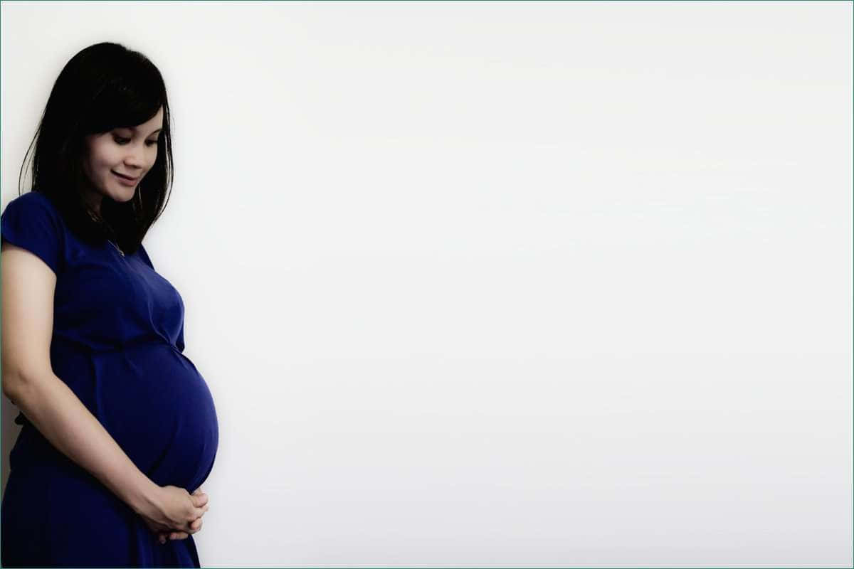 pregnancy 桌布 壁紙 — 一位身穿藍色連衣裙的孕婦，在純白背景前輕柔地抱著肚子，露出溫柔的微笑。