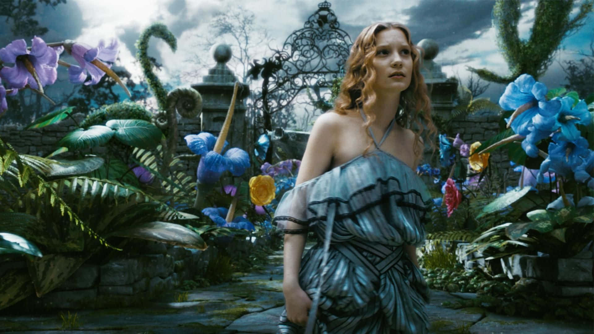 Mia Wasikowska 桌布 壁紙 — 一位身穿藍色長裙的女子，在陰天之下，置身於一個奇幻花園中，周圍是超大的彩色花朵和華麗的大門。