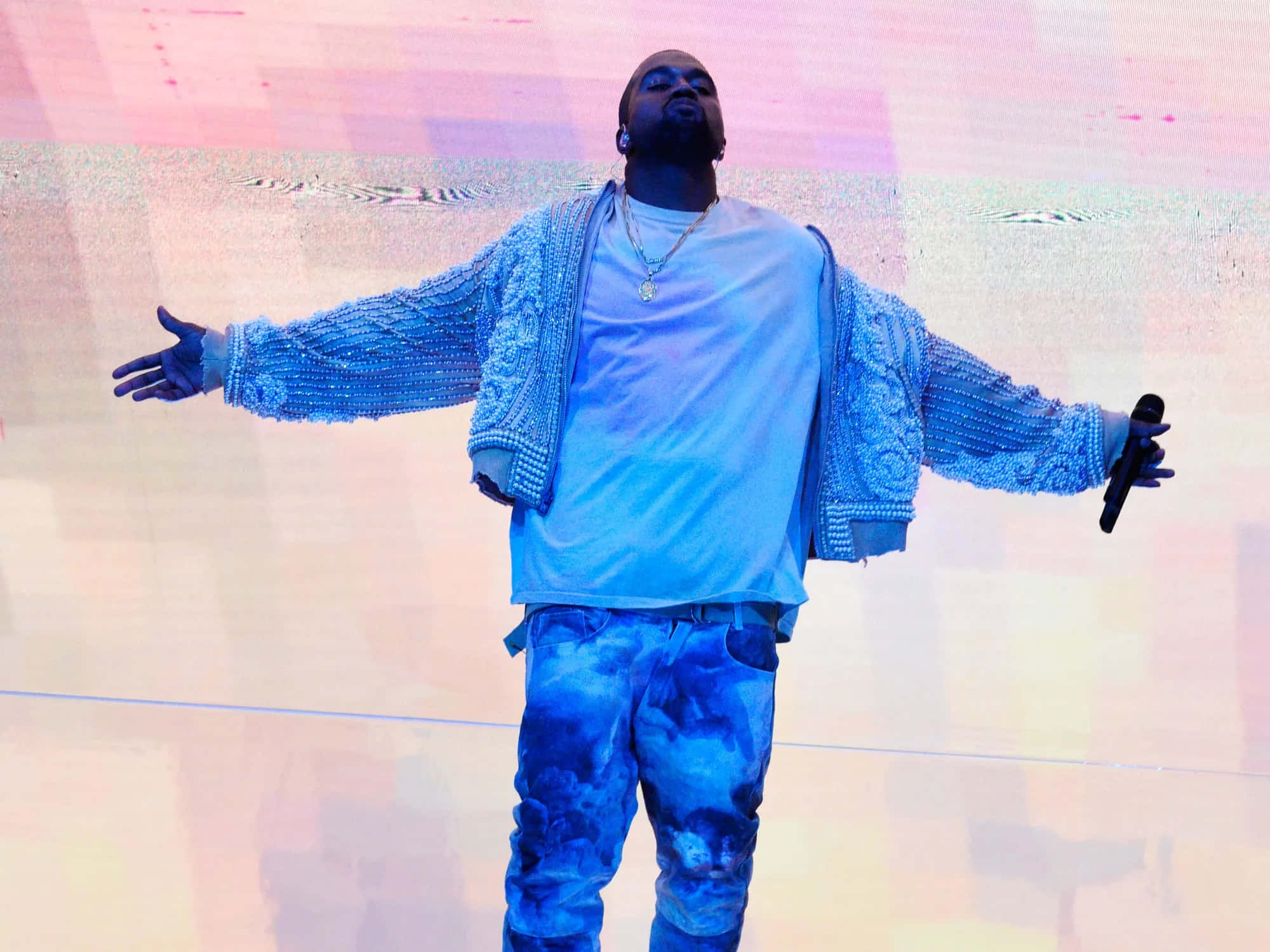 Kanye 桌布 壁紙 — 一位身穿藍色閃亮外套和紮染褲的表演者，在色彩繽紛的舞台上，在充滿活力的燈光下張開雙臂站立。