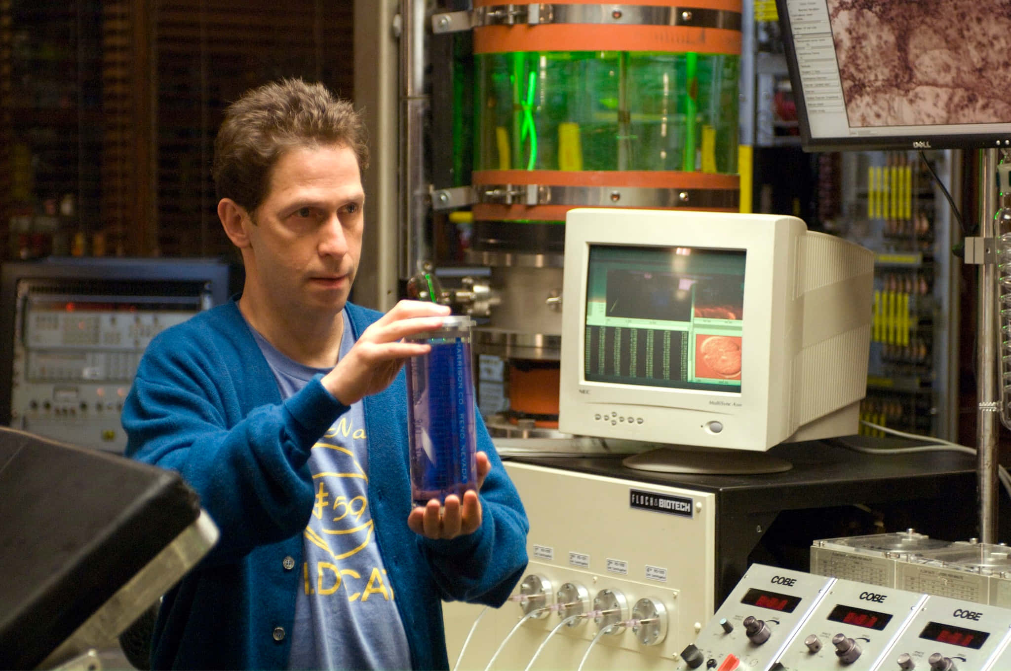Tim Blake Nelson 桌布 壁紙 — 一位身穿藍色開襟毛衣的科學家在充滿設備和顯示器的高科技實驗室中手持發光藍色小瓶。