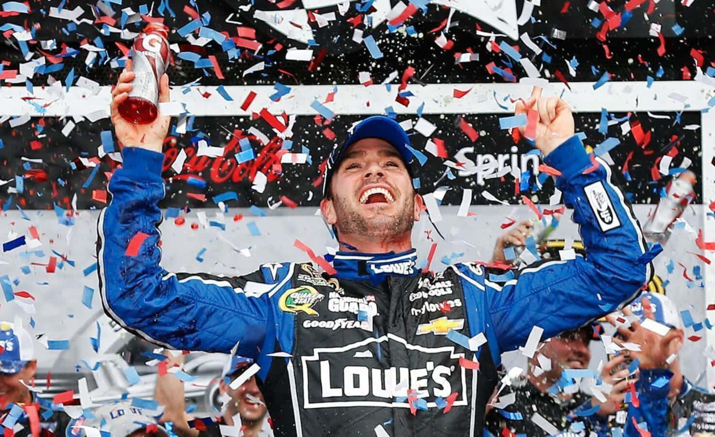 Jimmie Johnson 桌布 壁紙 — 一位身穿藍黑色賽車服的NASCAR車手高舉雙臂慶祝勝利，紅色、白色和藍色的五彩紙屑如雨般灑落在他周圍。