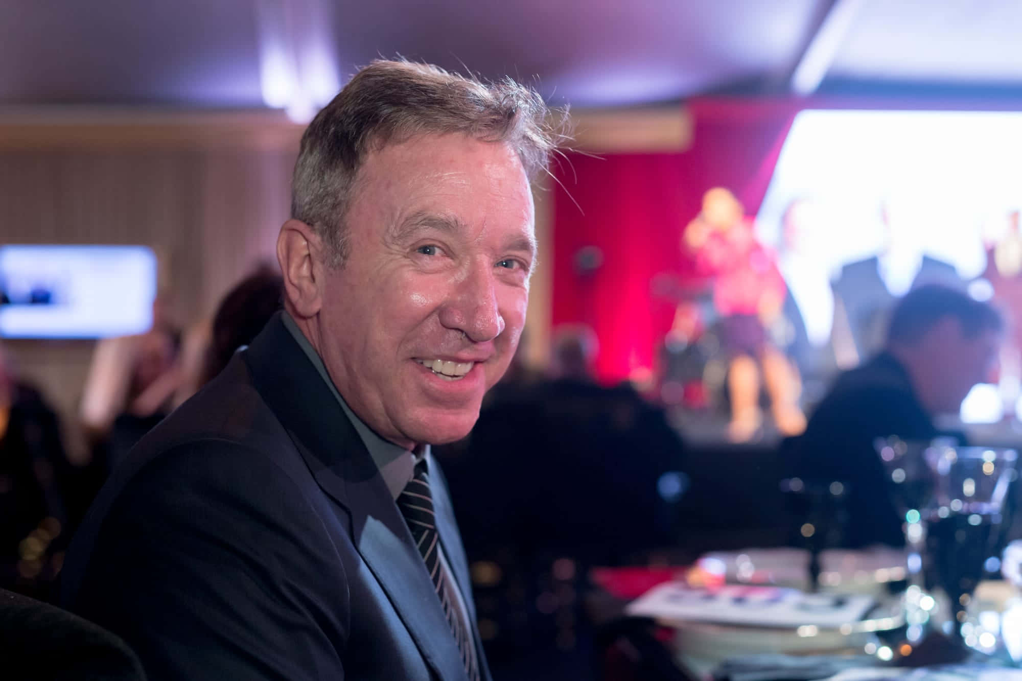 Tim Allen 桌布 壁紙 — 一位身穿西裝、面帶微笑的男子在一個有舞台和紅色燈光的熱鬧活動中望向鏡頭。