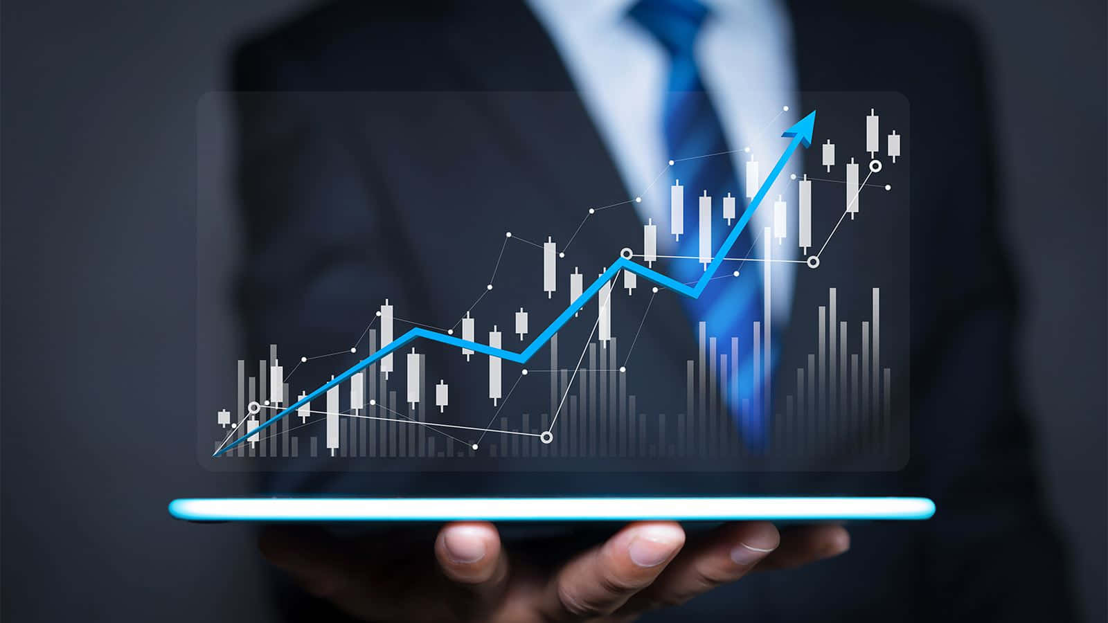 Trading 桌布 壁紙 — 一位身穿西裝的商人手持發光平板電腦，顯示帶有蠟燭圖形態的上升藍色股票圖表。