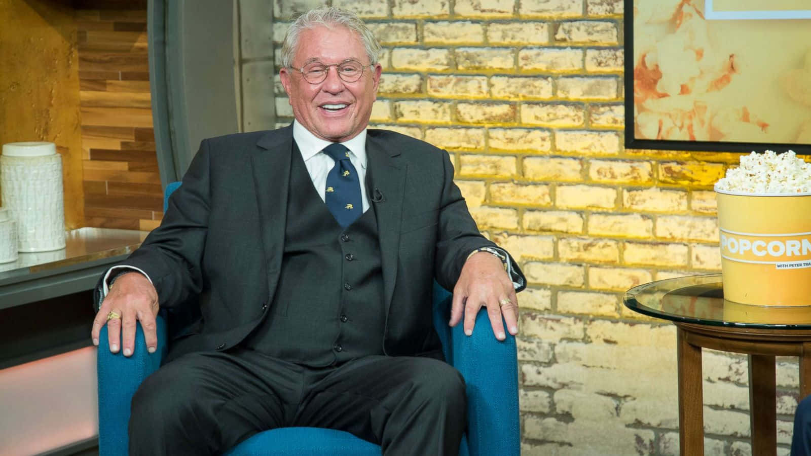 Tom Berenger 桌布 壁紙 — 一位身穿西裝的年長男士坐在藍色扶手椅上，旁邊放著一桶爆谷，開懷大笑。