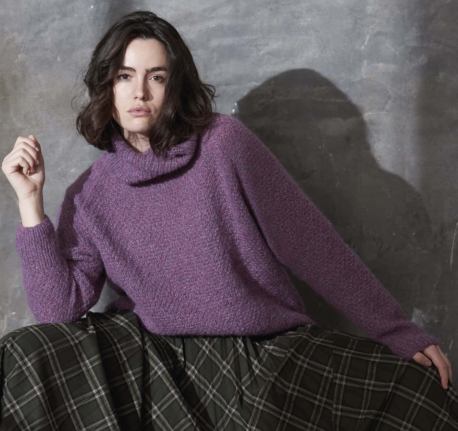 Purple Turtle-neck Sweater 桌布 壁紙 — 一位身穿質感紫色高領毛衣和格子裙的女性，在灰色混凝土牆前擺姿勢，帶有戲劇性的陰影。