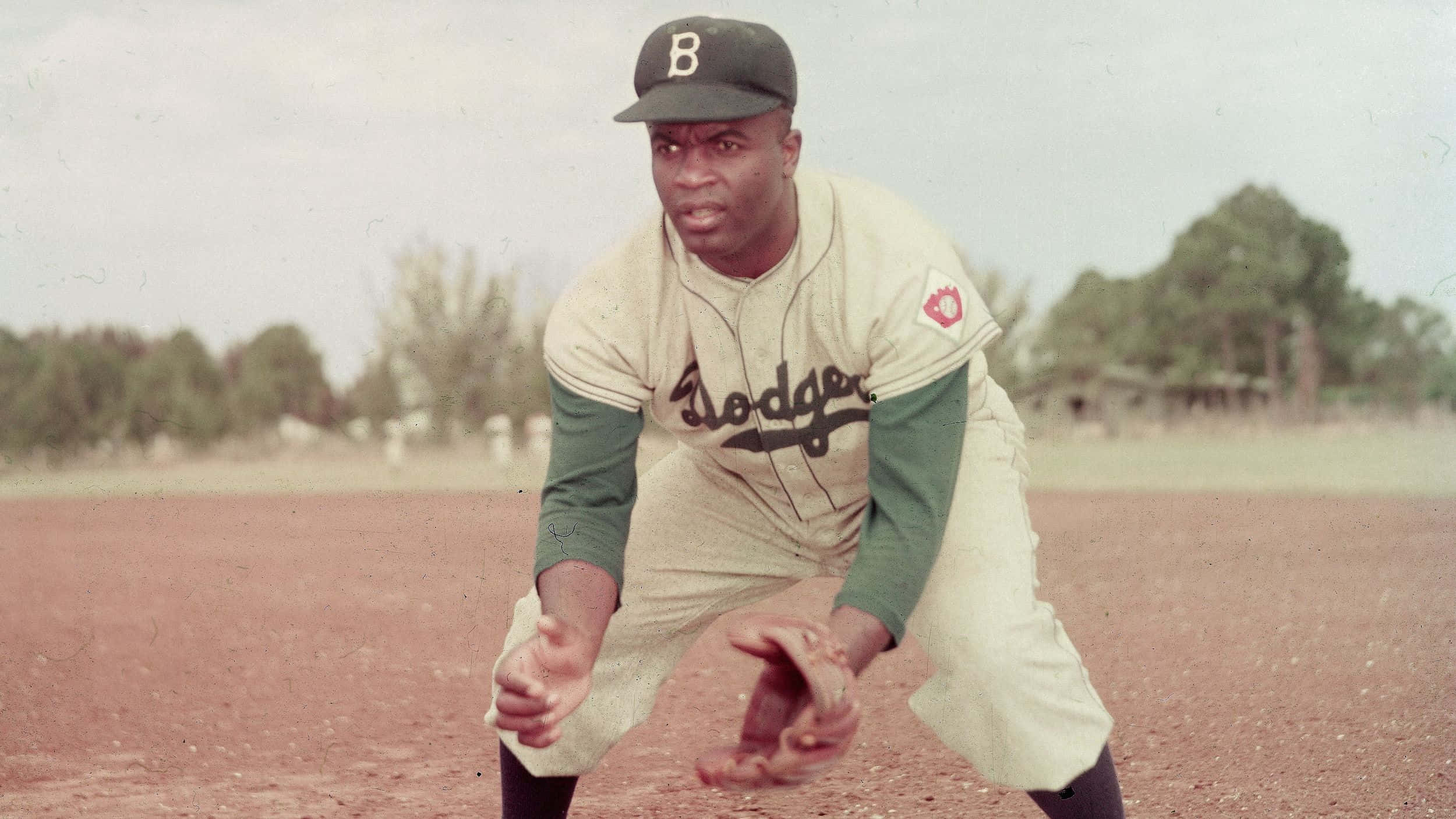 Jackie Robinson 桌布 壁紙 — 一位身穿道奇隊球衣的復古棒球運動員在陽光照耀的泥土場地上蹲伏準備，背景有樹木。