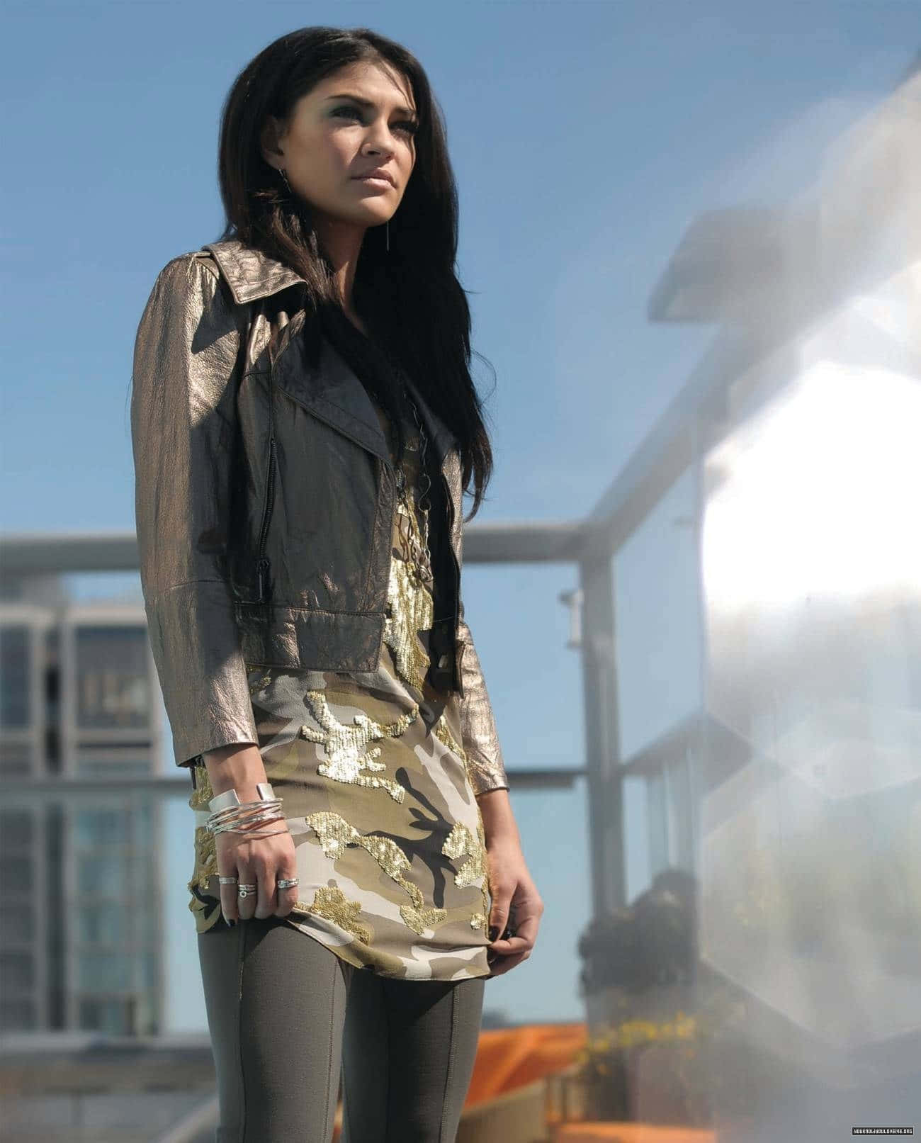 jessica szohr 桌布 壁紙 — 一位身穿金屬色外套和圖案連衣裙的女性，在明亮的藍天和現代建築背景下自信地站立。