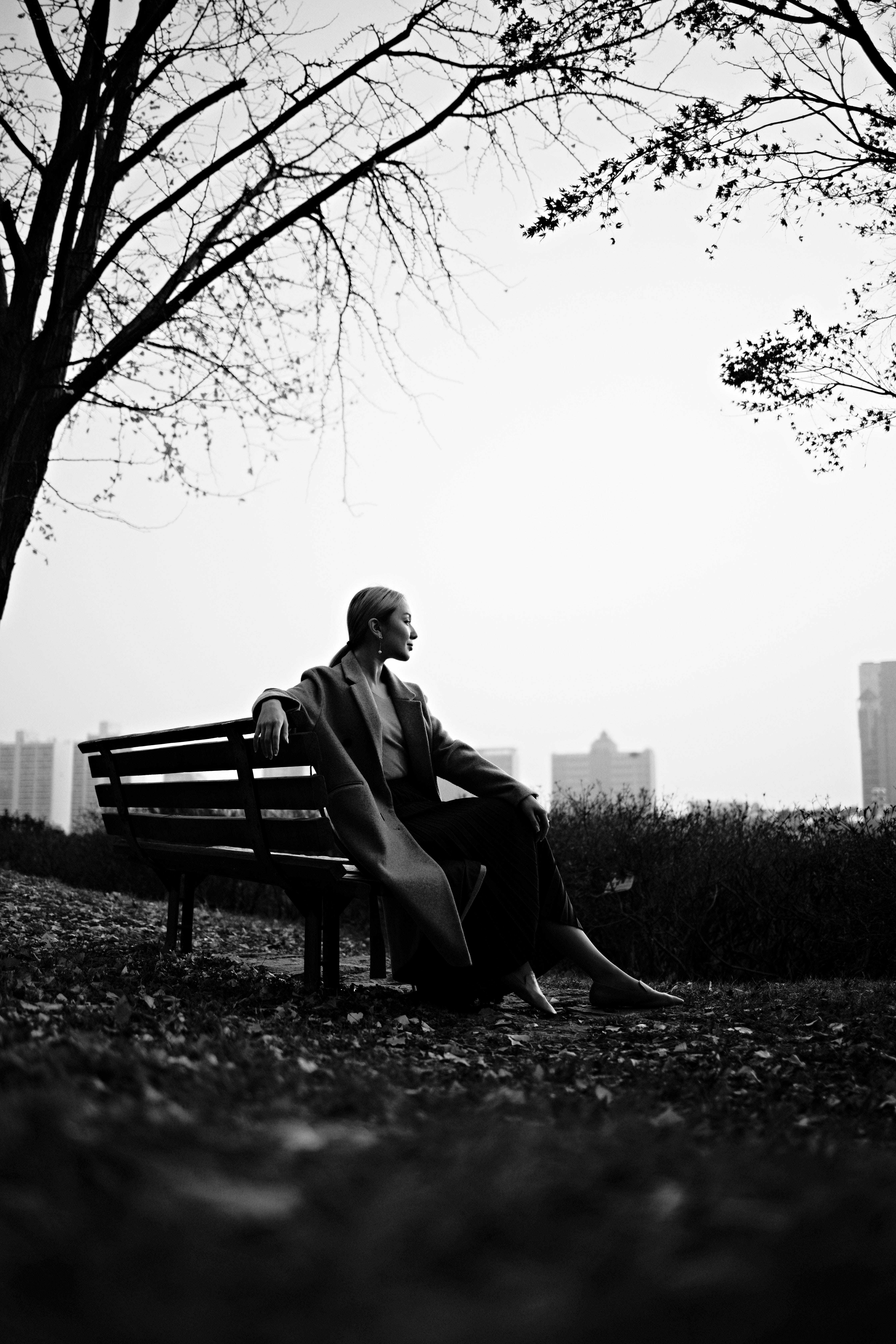 Sitting Alone 桌布 壁紙 — 一位身穿長外套的女性獨自坐在公園長椅上，頭頂是光禿禿的樹枝，凝視著朦朧的城市天際線，呈現單色調。