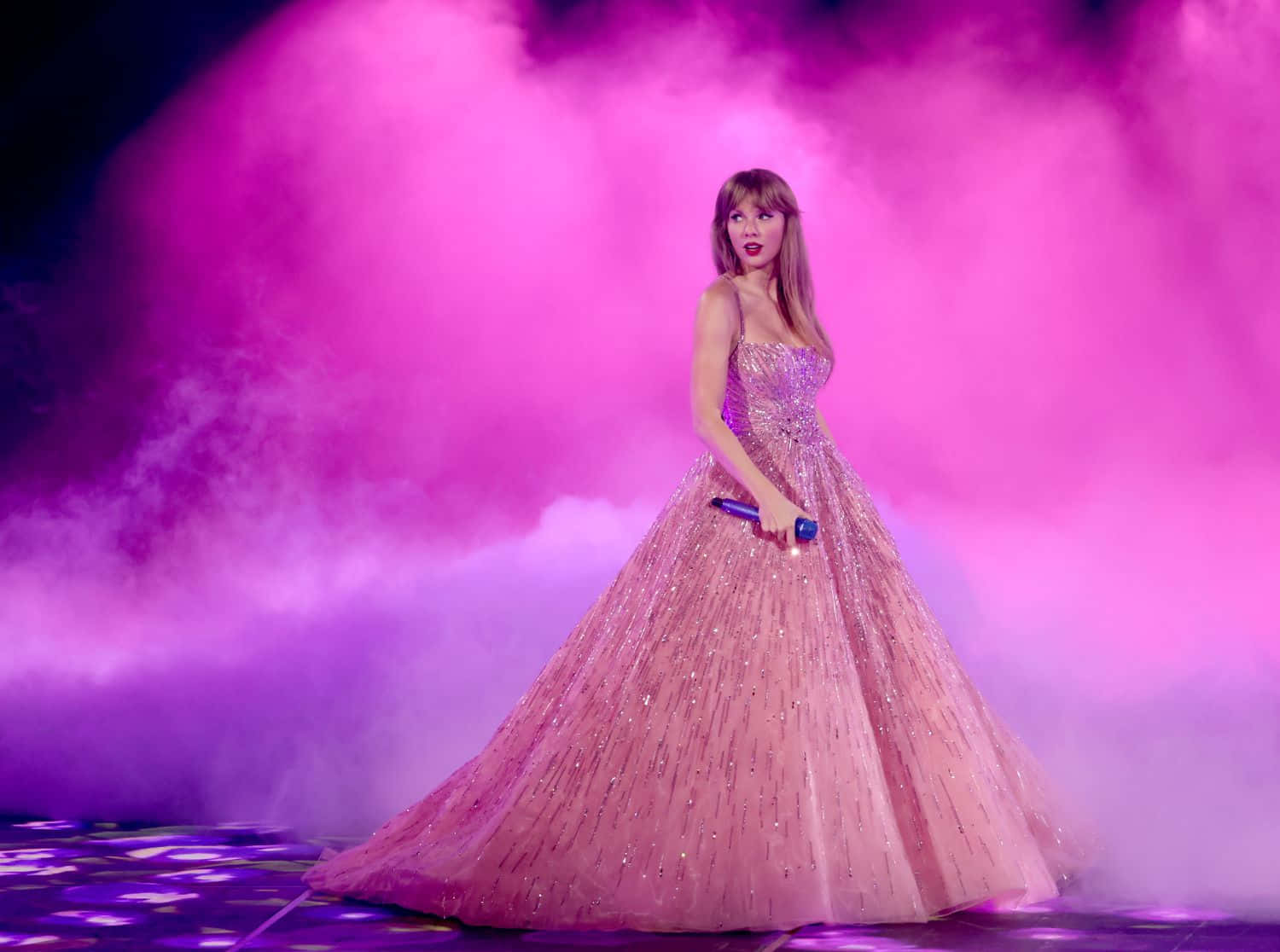 Taylor Swift 桌布 壁紙 — 一位身穿閃亮粉紅色禮服的歌手站在舞台中央，沐浴在鮮豔的粉紅色煙霧和紫色燈光中。
