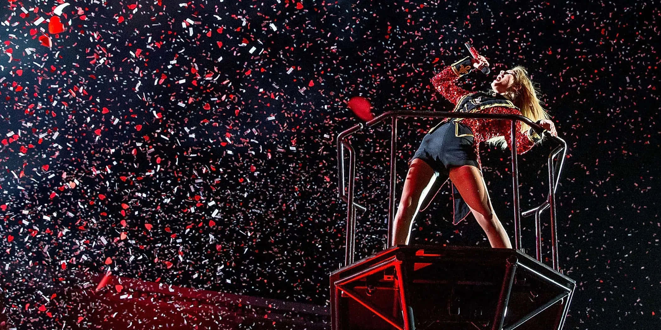 Taylor Swift 桌布 壁紙 — 一位身穿閃亮紅色服裝的女歌手在高台上熱情表演，紅色和白色的紙屑在她周圍爆炸，背景是黑暗的舞台。