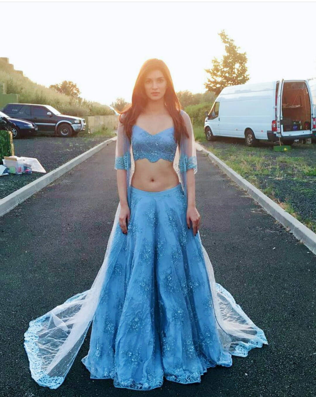 連身裙 桌布 壁紙 — 一位身穿閃亮藍色 lehenga 的女性站在瀝青路上，正值黃金時段，附近停有車輛。