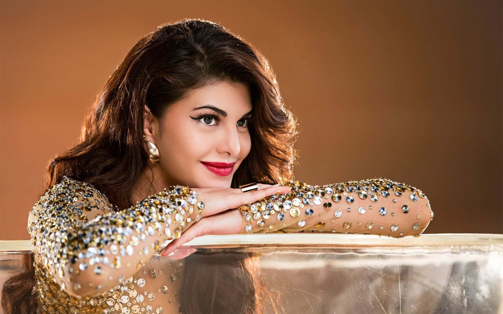Jacqueline Fernandez 桌布 壁紙 — 一位身穿閃亮金色亮片上衣的迷人女士，在溫暖的棕色背景下優雅地靠在玻璃表面上。