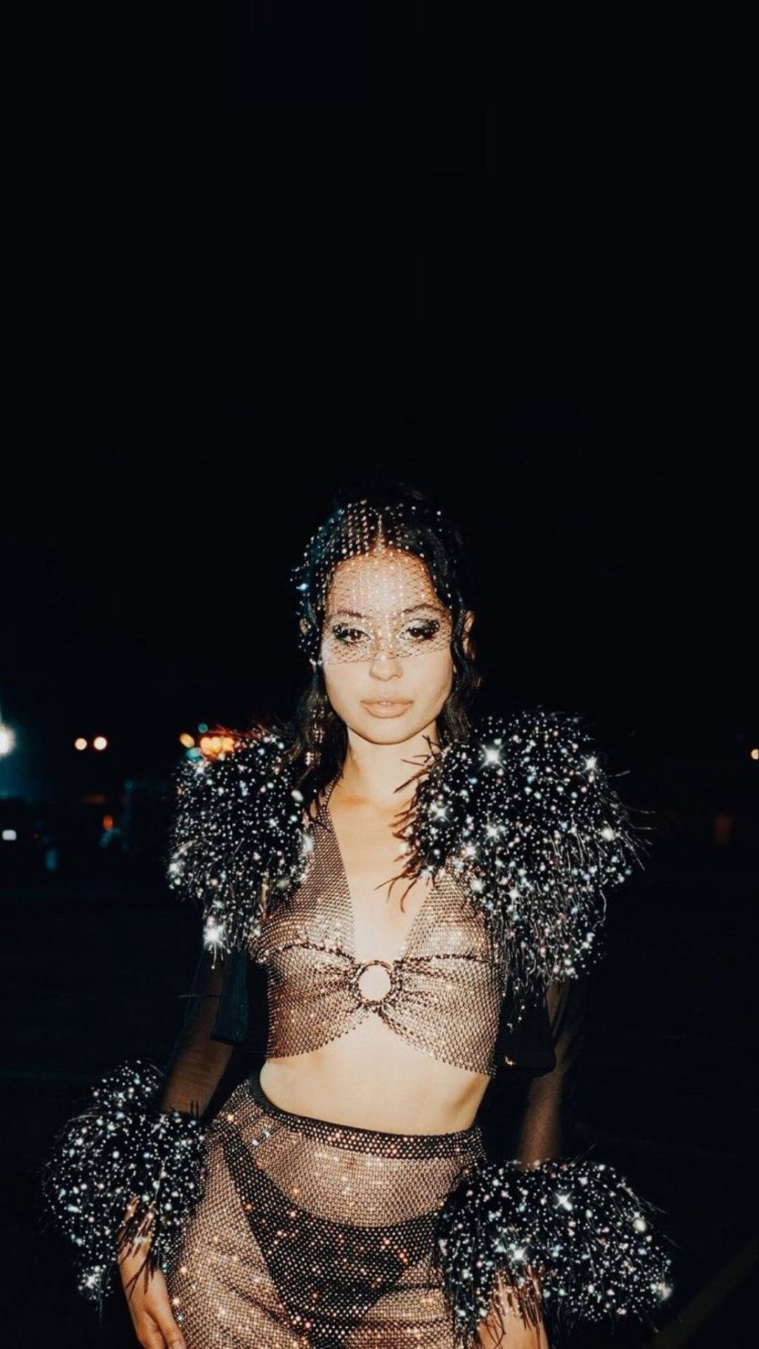 Euphoria Maddy Perez 壁紙 — 一位身穿閃亮銀色亮片服裝、帶有泡泡袖的女性，在黑暗的夜晚背景下自信地站立，遠處有城市燈光。