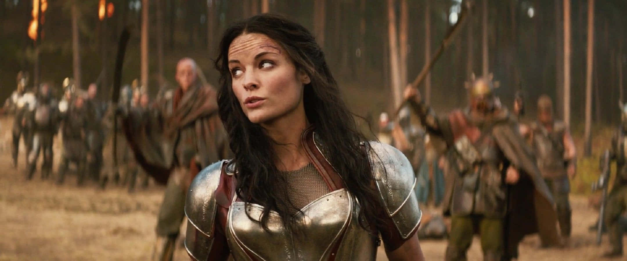 Lady Sif 桌布 壁紙 — 一位身穿閃亮銀色盔甲的女戰士在陽光明媚的森林中警覺地站立，身後有模糊的士兵在行進。