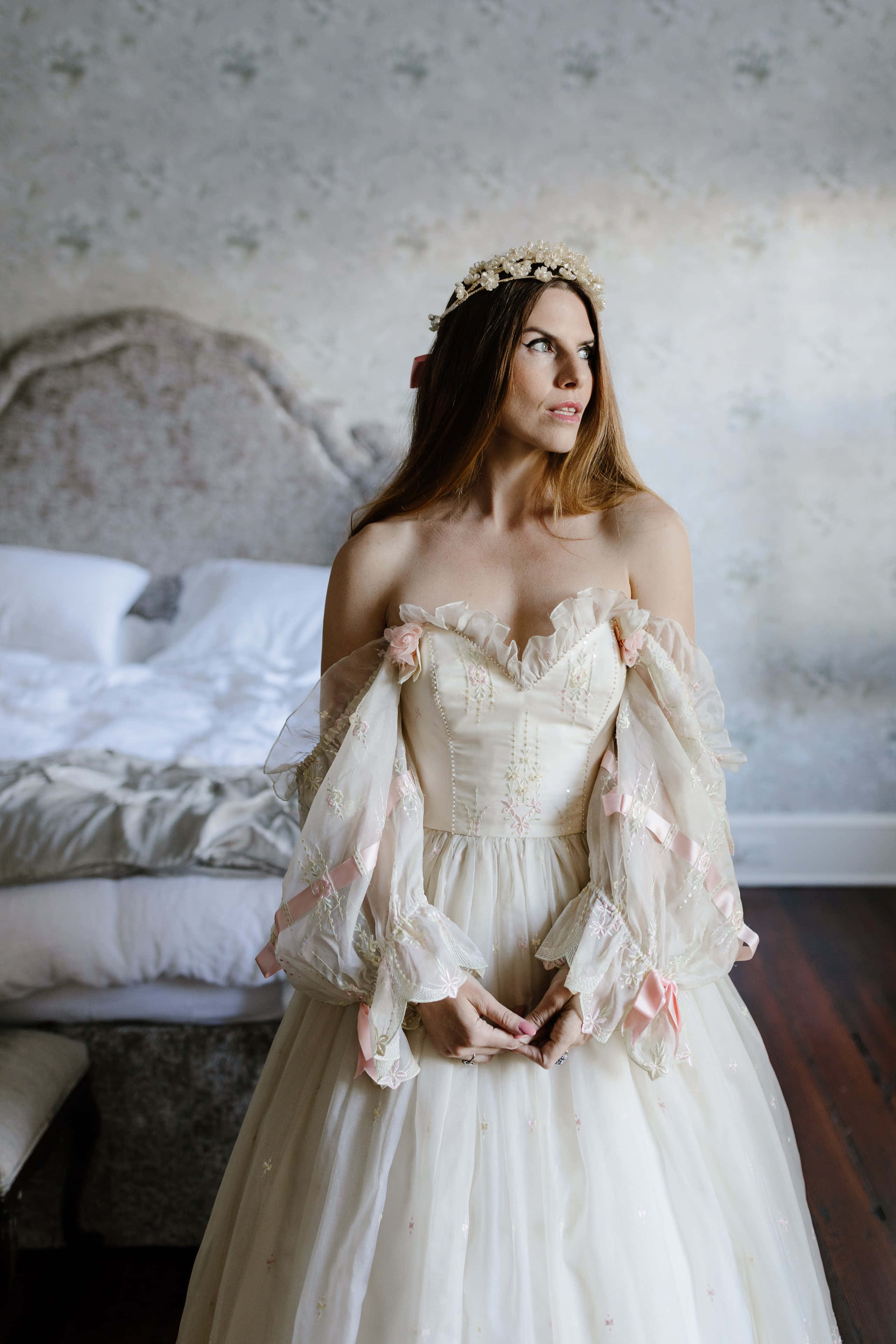 Bride's Dress 桌布 壁紙 — 一位身穿露肩象牙色禮服、繫著粉紅色絲帶的女性，站在燈光柔和、舖有花卉壁紙和復古床的臥室中。